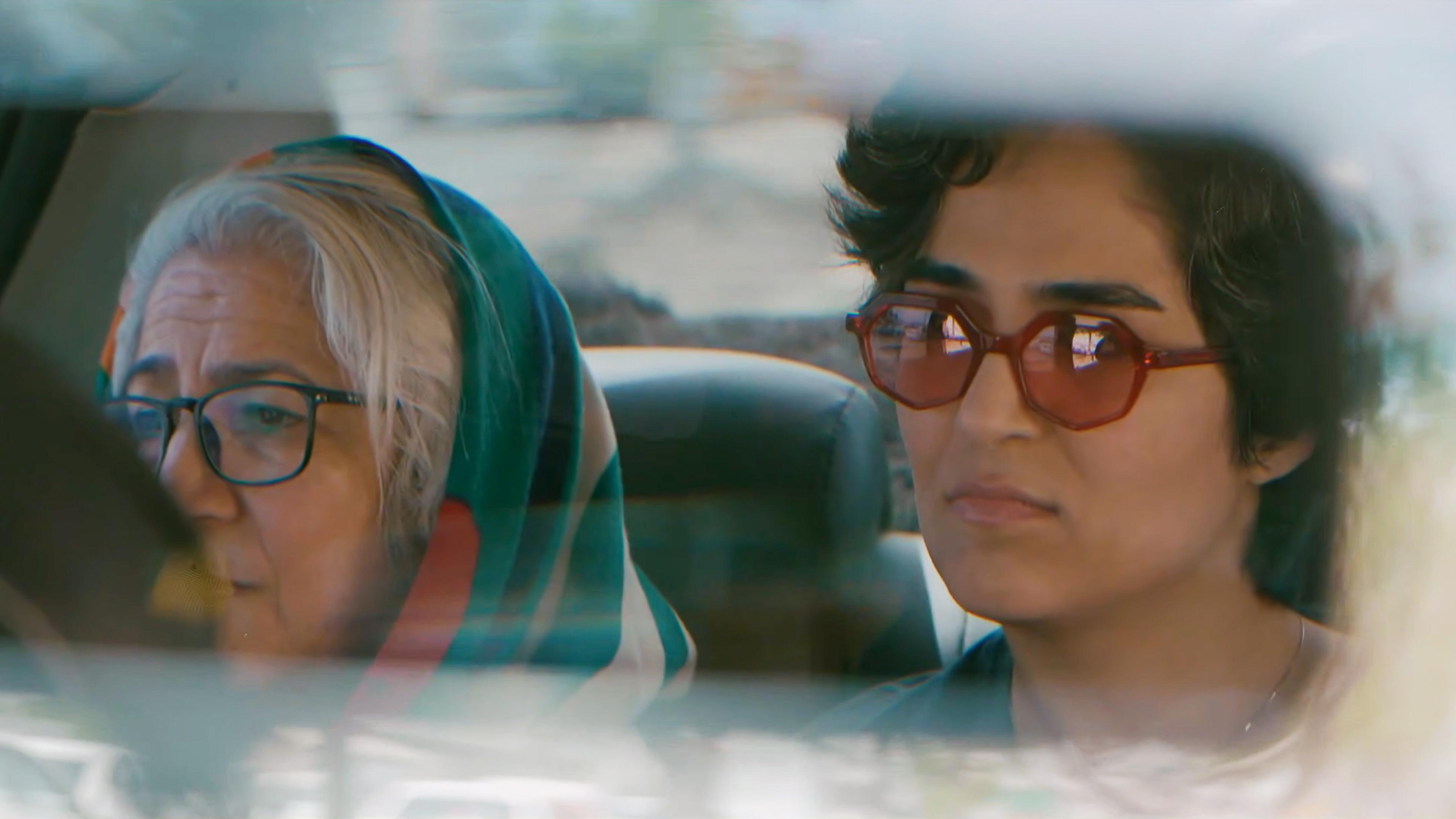 Two women sitting in a car, one is wearing glasses and a headscarf, the other has short hair and red sunglasses.