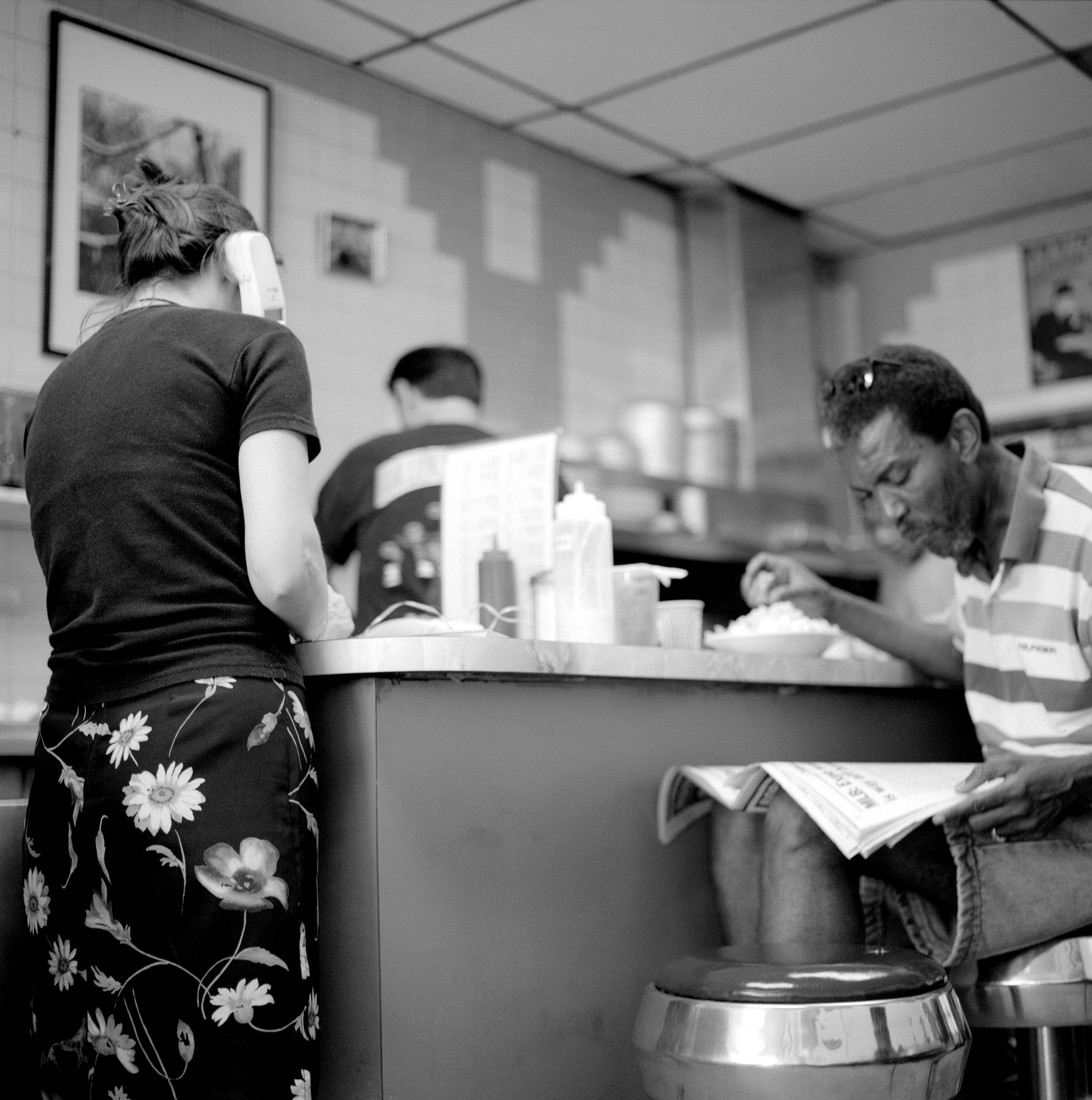 Black and white photo of a diner scene with a woman using a phone and a man reading a newspaper at a counter.