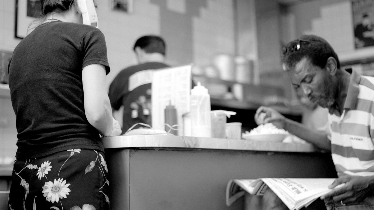 Black and white photo of a diner scene with a woman using a phone and a man reading a newspaper at a counter.