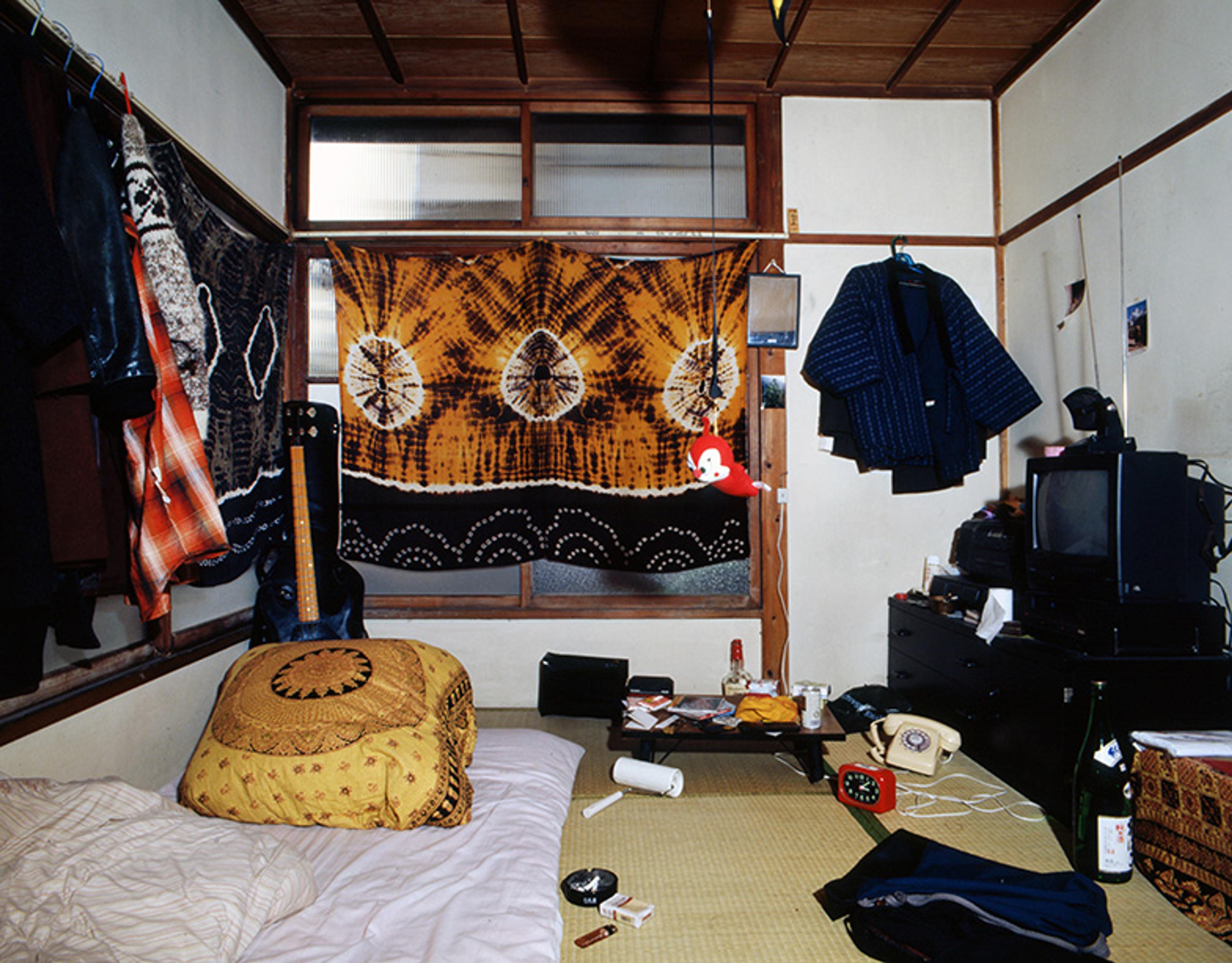 A bedroom with a futon, a tie-dyed textile covering the window and various items strewn on the floor, including a guitar, alcohol bottles, a cigarette pack and ashtray, a rotary phone and a paint roller.