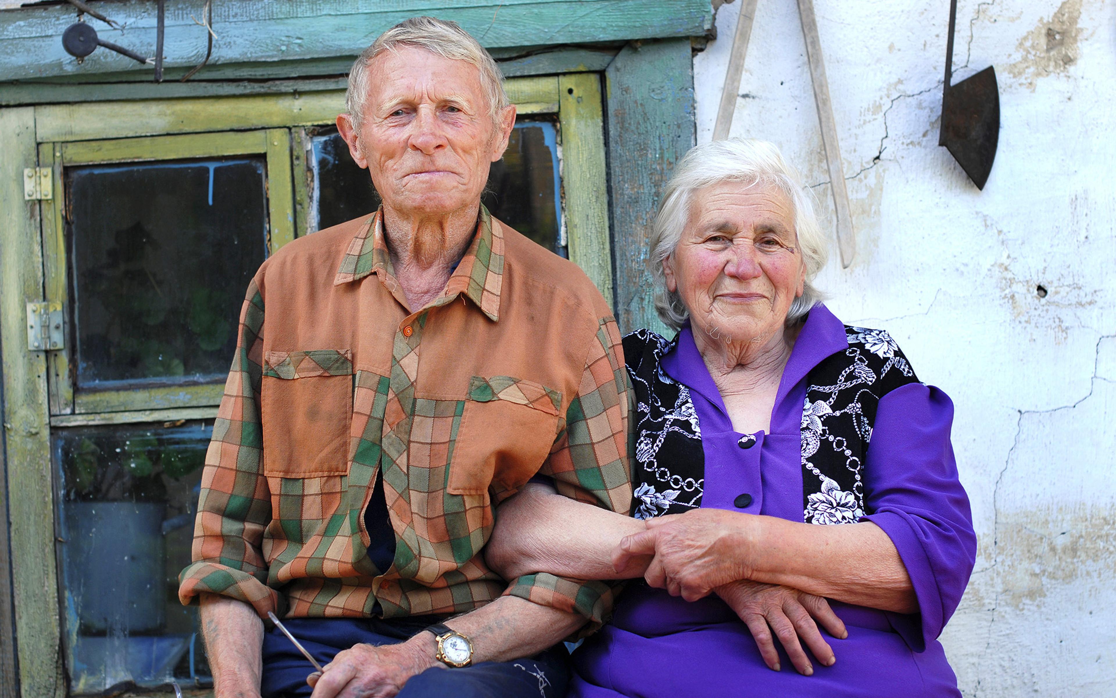 An elderly man and woman sitting together outside an old, rustic house, the man wears a plaid shirt, the woman a purple dress. They look happy.