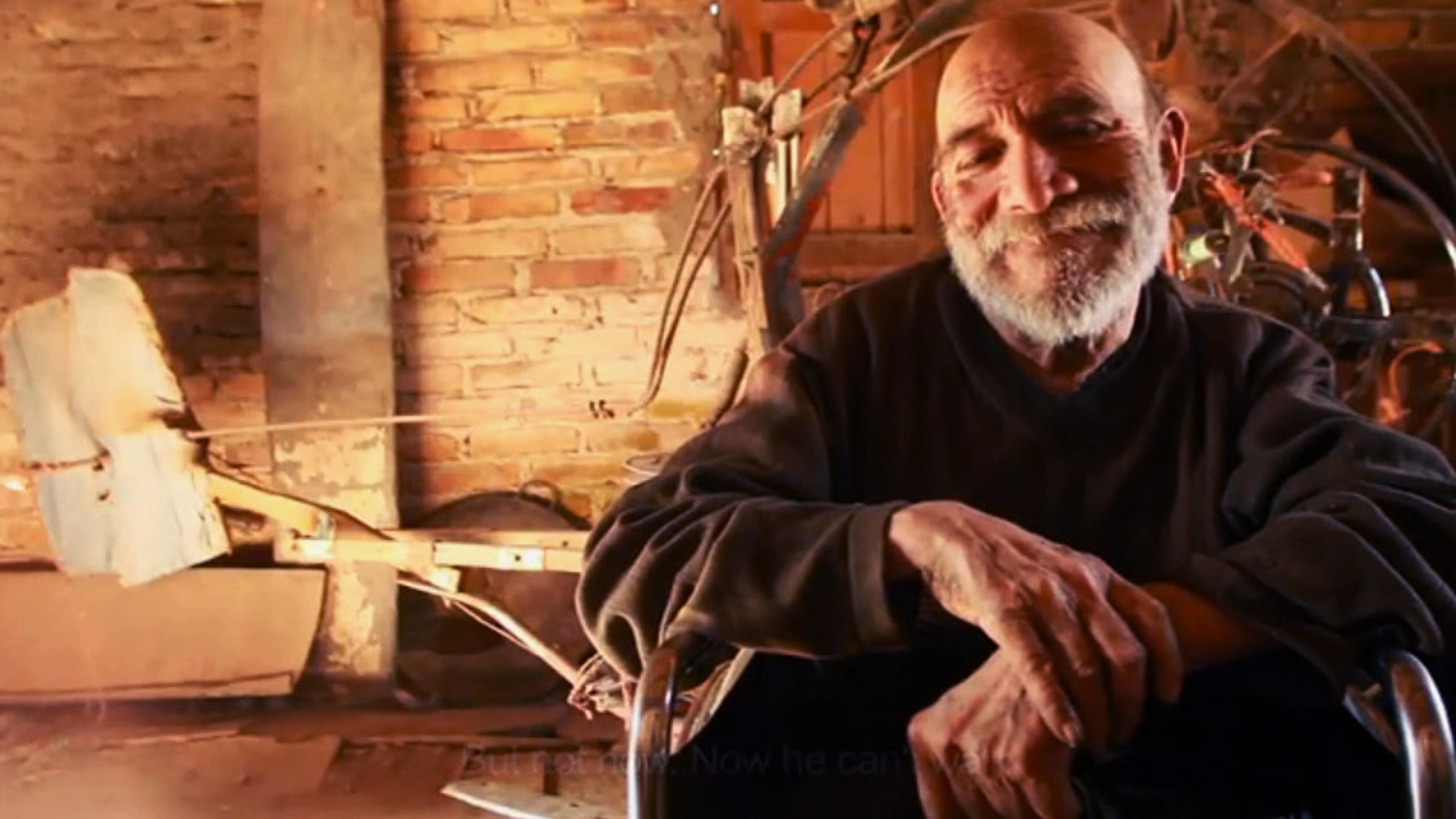 An elderly man with a beard smiling, sitting indoors against a brick wall with wooden objects and mechanical parts.