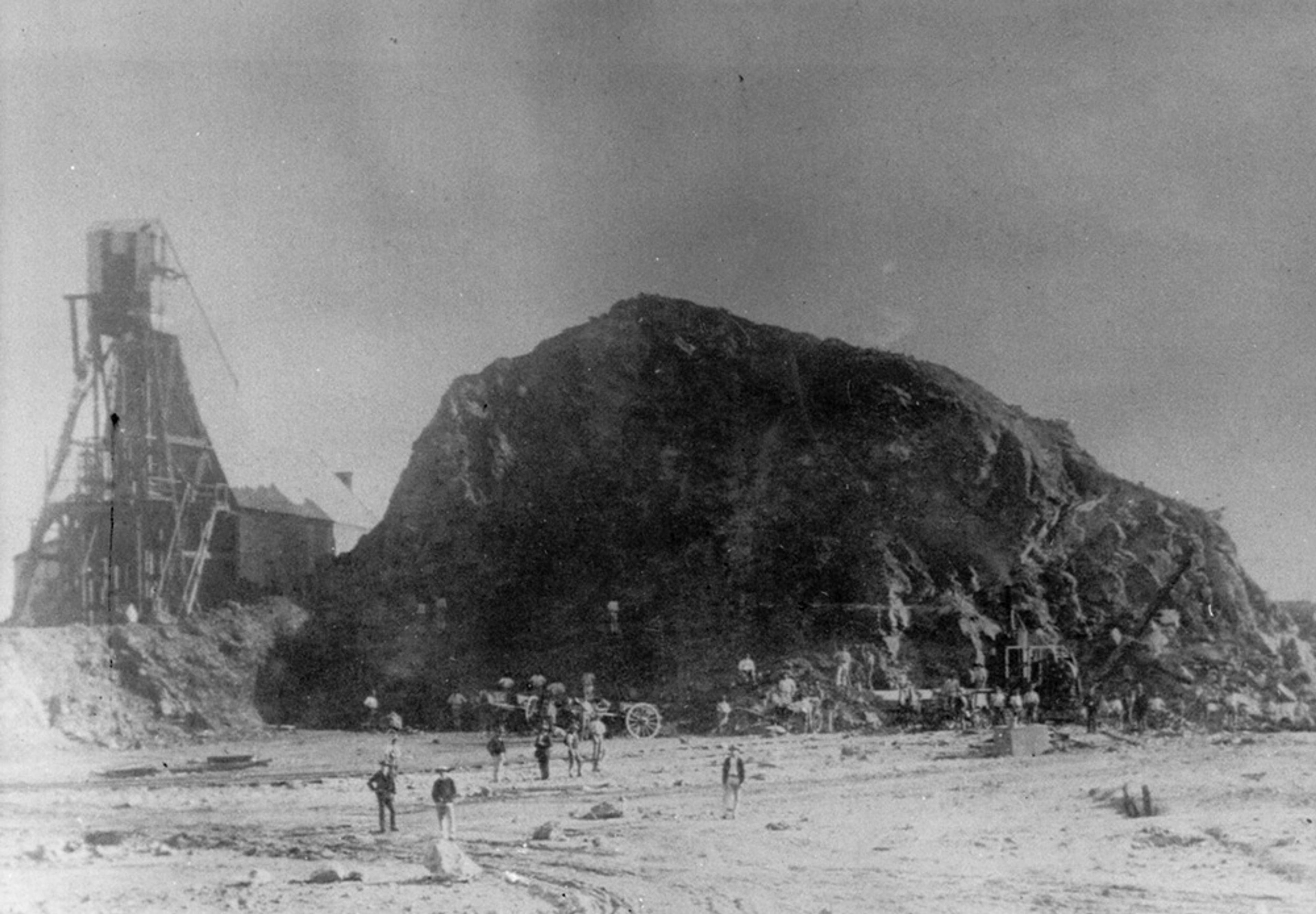 Black and white photo of an old mining site with a large rock pile, a wooden mining structure and several people in the foreground.