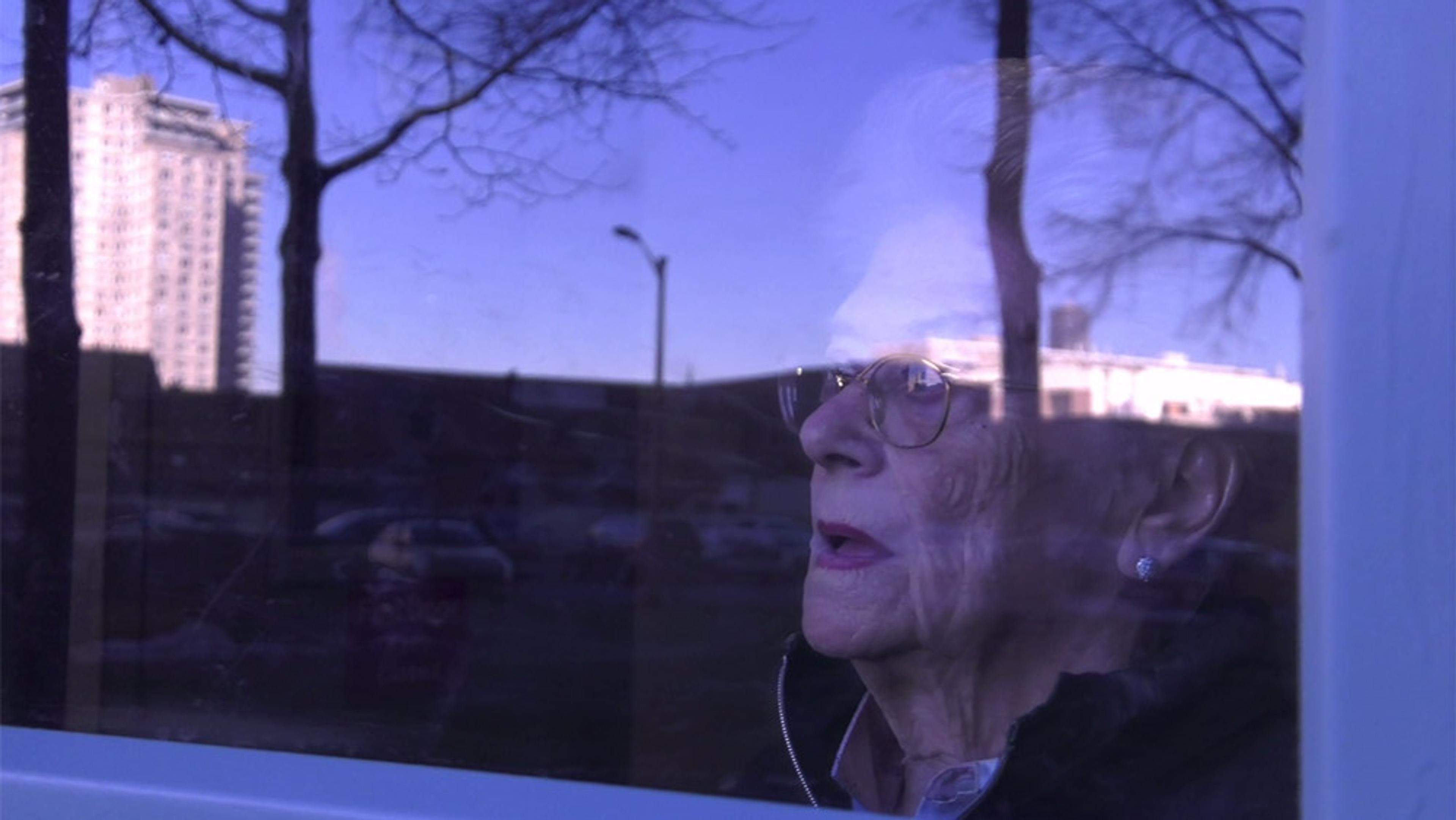 An elderly woman looking out a window with reflections of trees and a building under a blue sky.