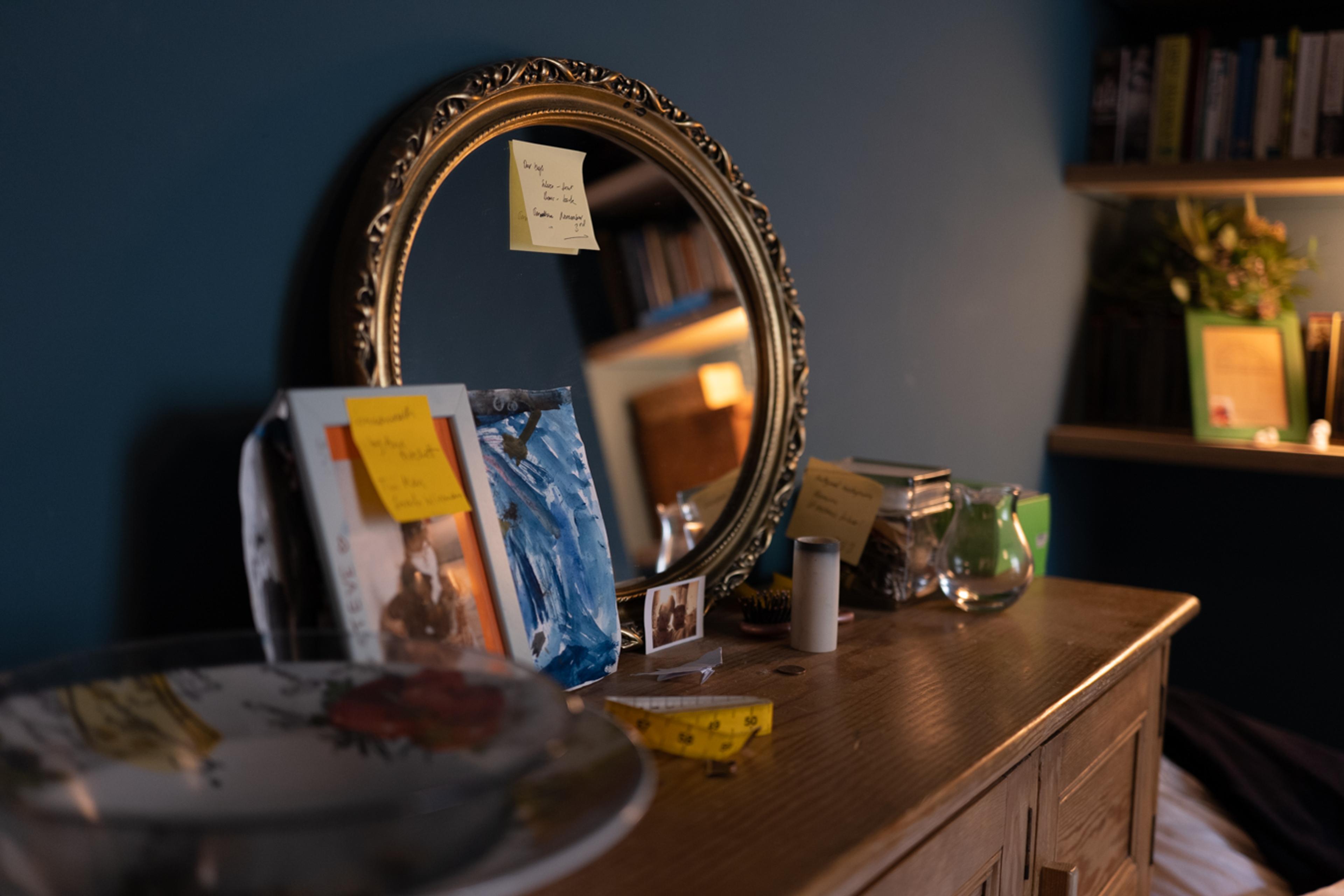 Photo of a wooden dresser with a round mirror books and notes. A tape measure and candle holder are on the dresser.