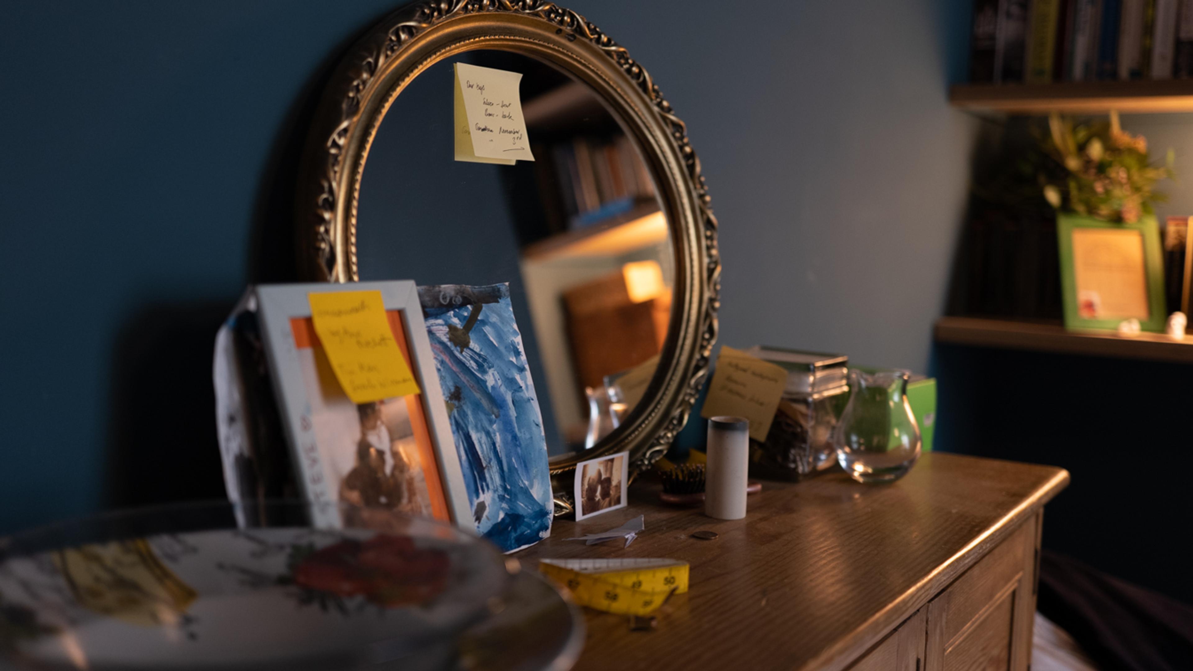 Photo of a wooden dresser with a round mirror books and notes. A tape measure and candle holder are on the dresser.