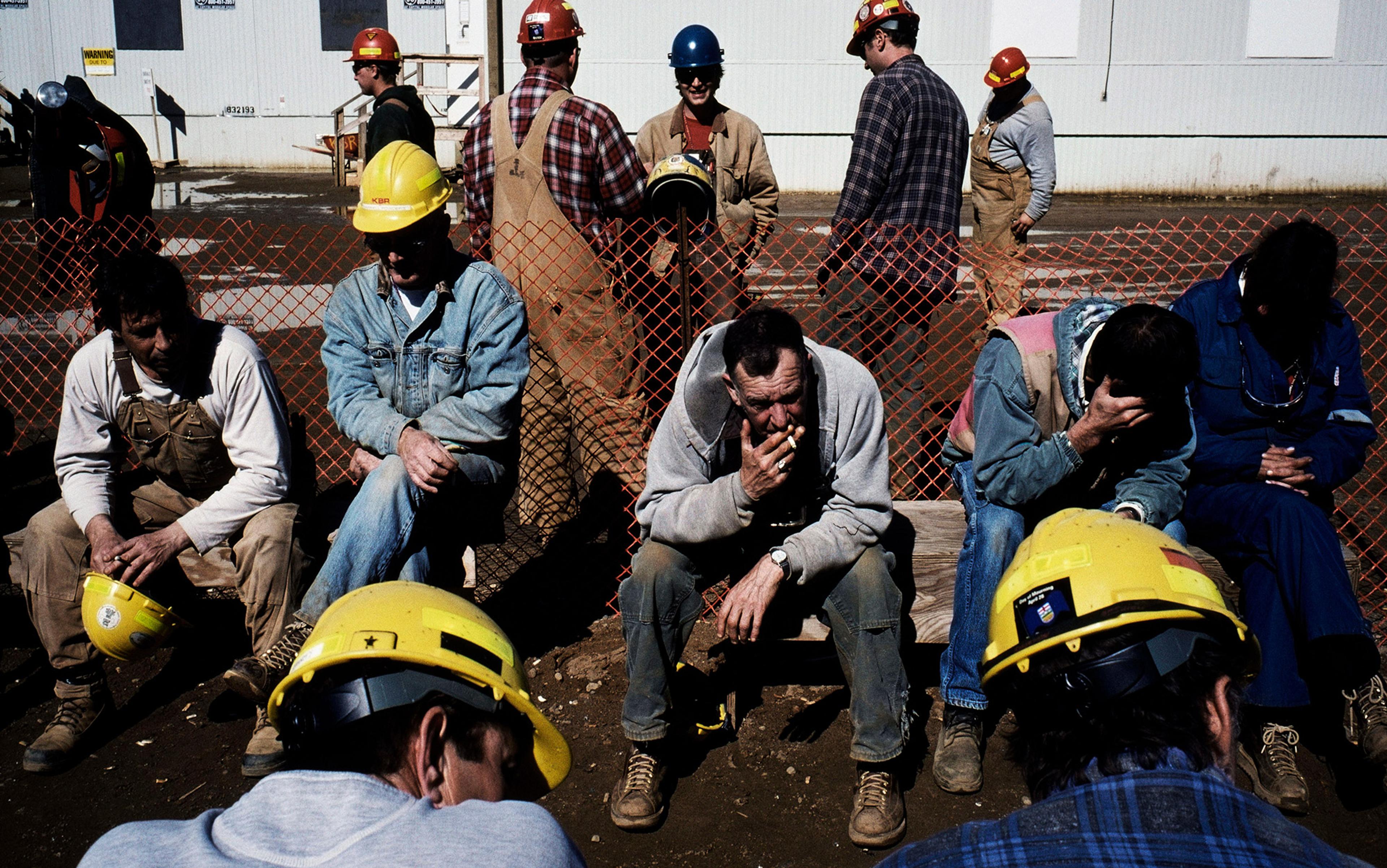 Construction workers sitting and standing around, some wearing hard hats, during a break outside a building, with a temporary orange safety fence visible.