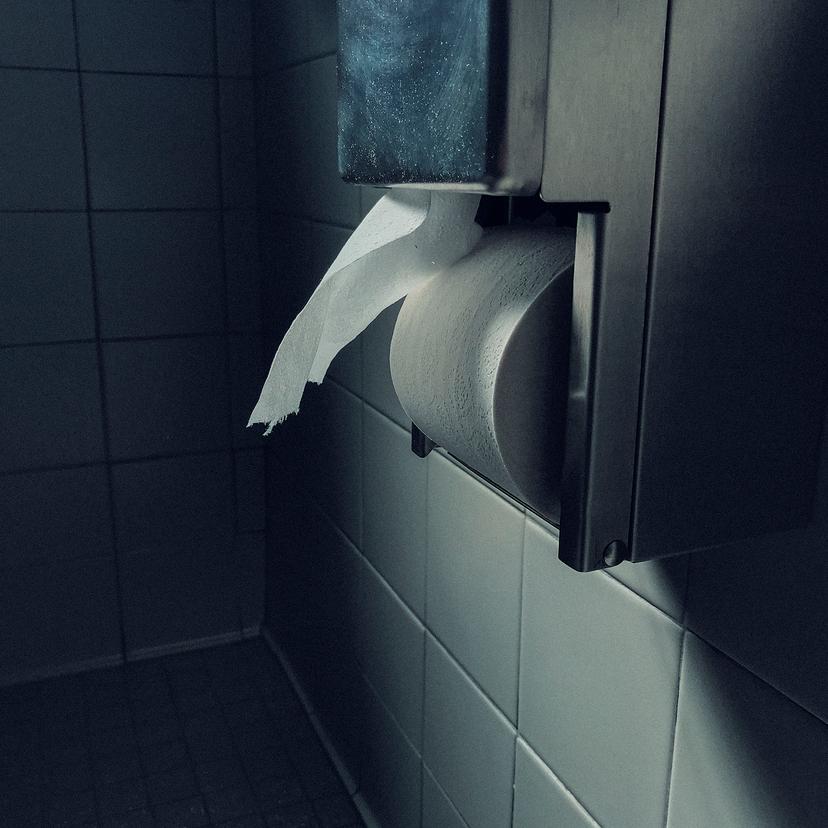 A bathroom scene featuring a metal toilet roll dispenser with a partially unrolled toilet paper, mounted on a tiled wall.