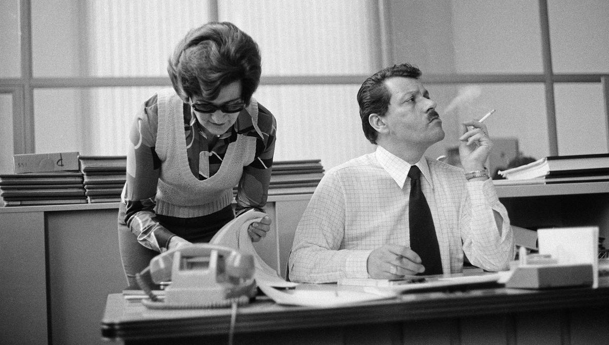 Two office workers, one standing and reviewing papers, the other seated and smoking, in a 1970s-style office with a telephone and stacks of paperwork.