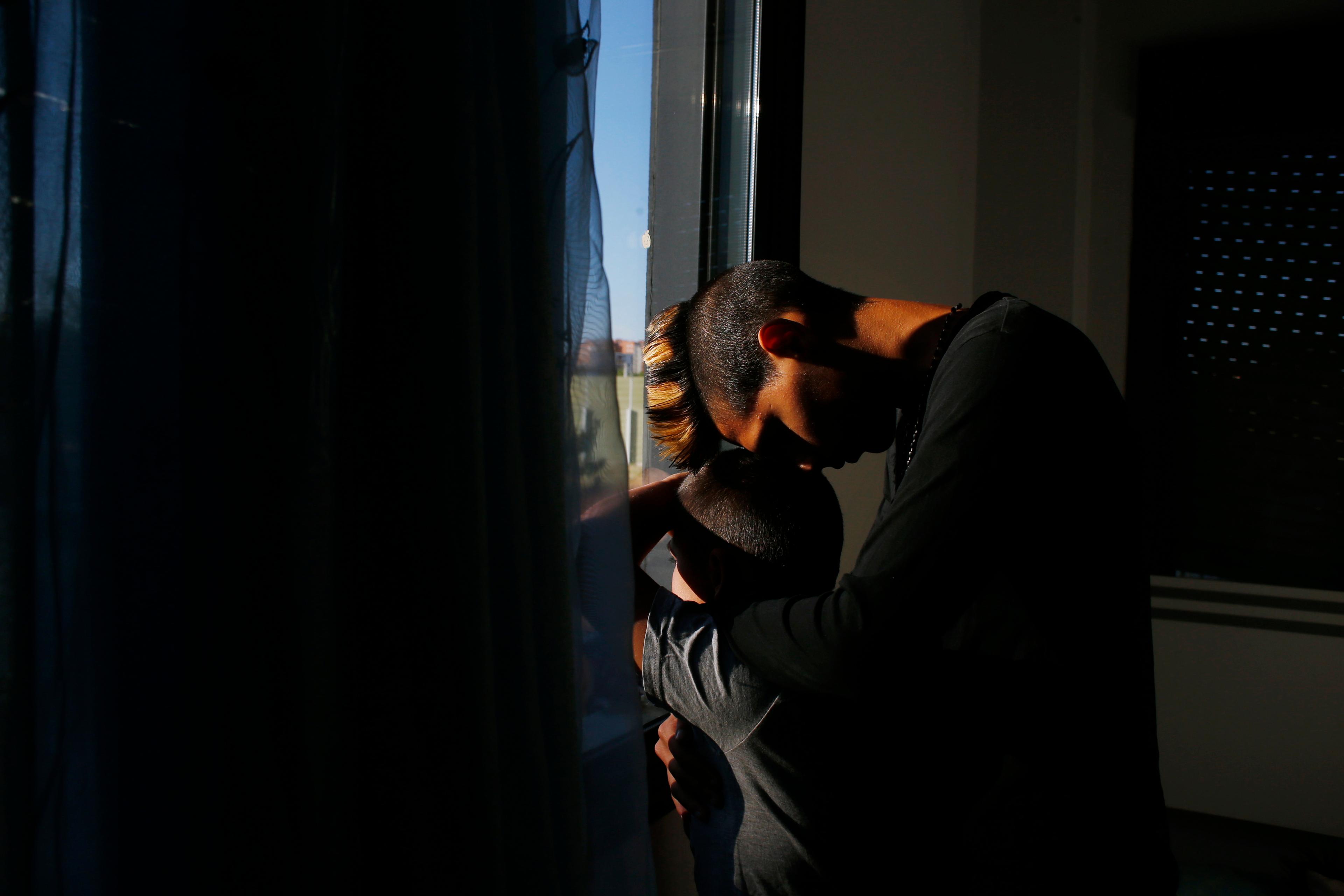 Photo of two people embracing by a window, with dramatic lighting creating shadows on their faces and the wall.