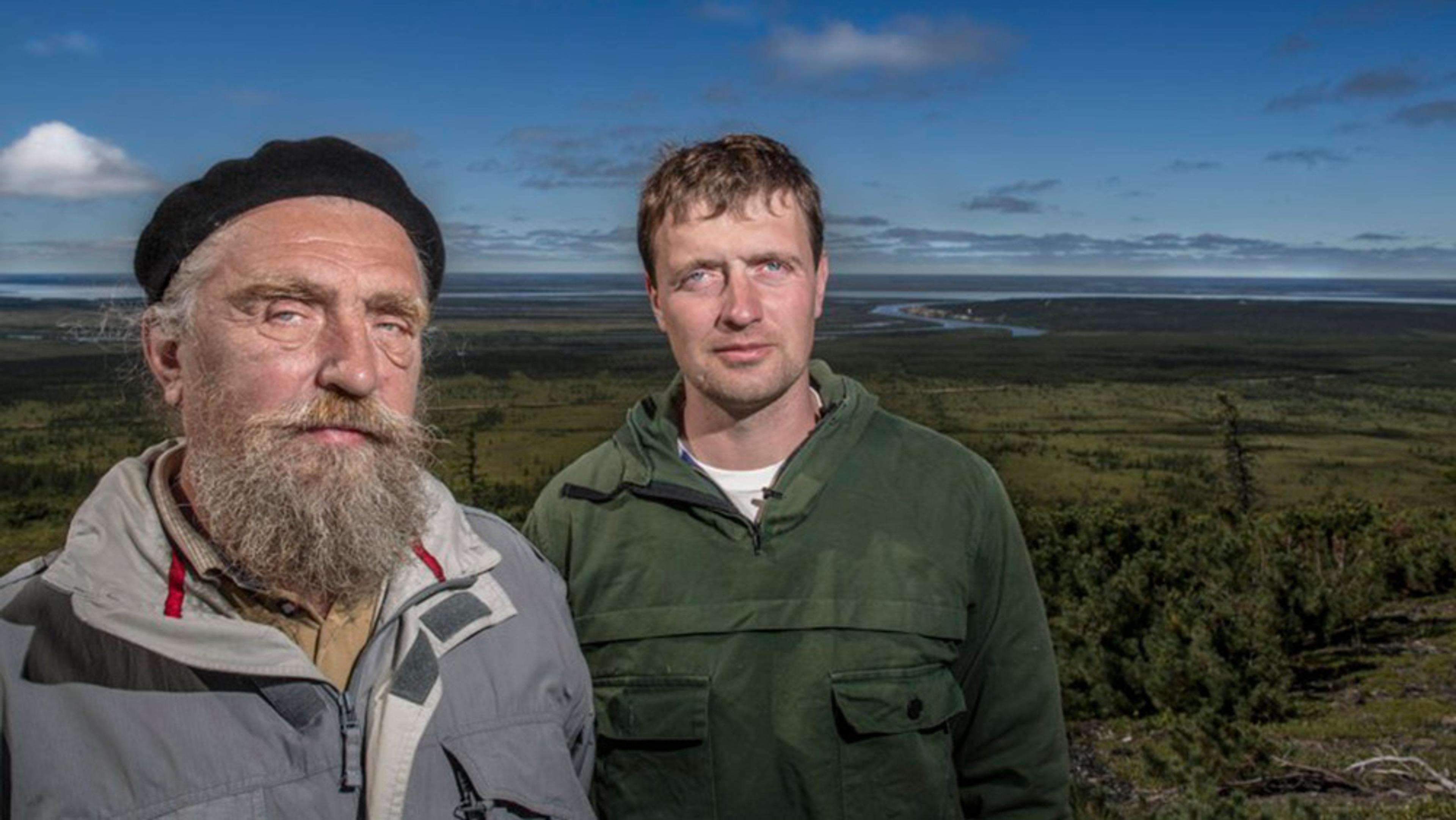 Two men outdoors with a vast green landscape and blue sky in the background, the older, beaded one wearing a cap and the other in a green jacket.