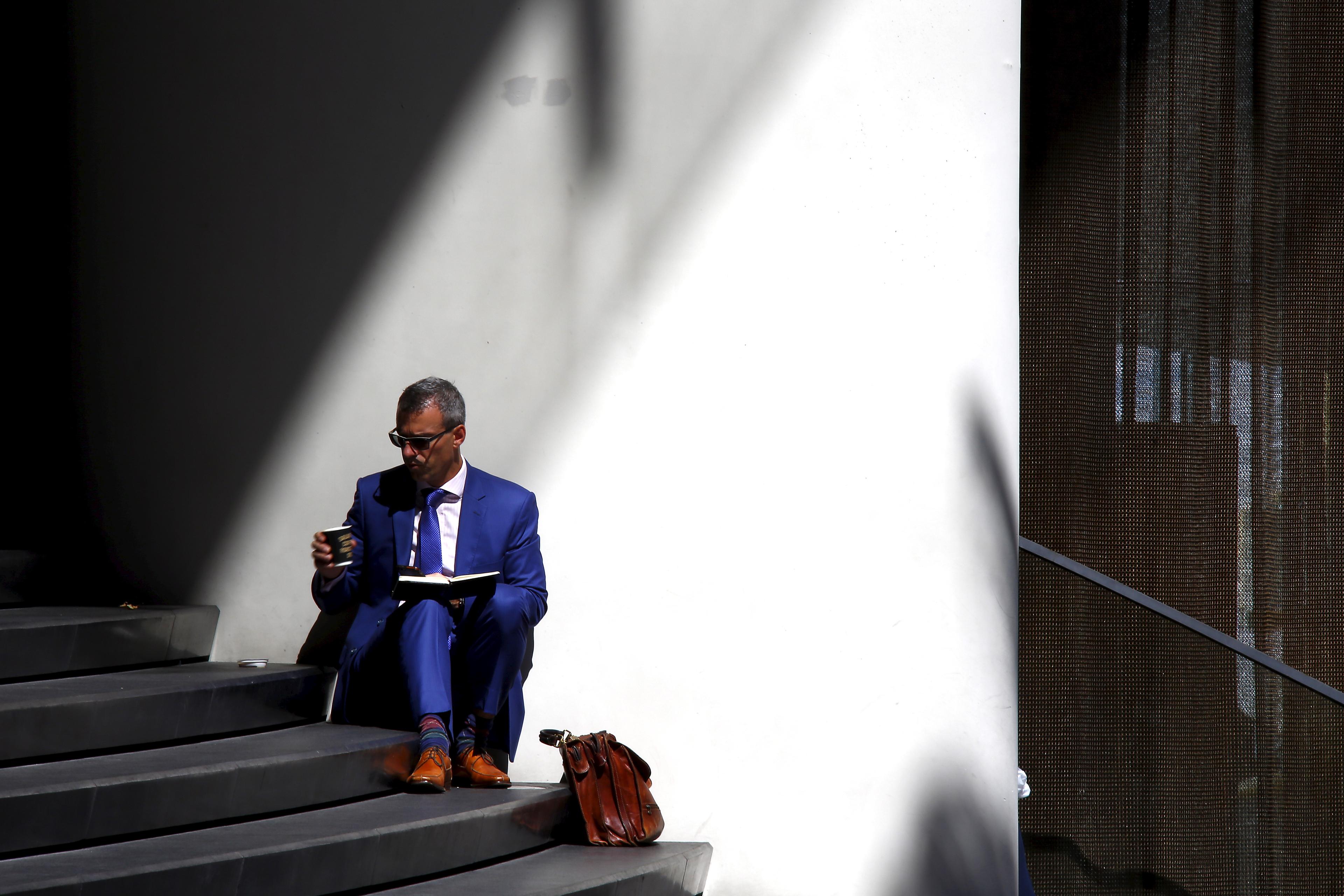 Photo of a man in a blue suit sitting on steps, reading a book, holding a coffee cup with a brown bag beside him.