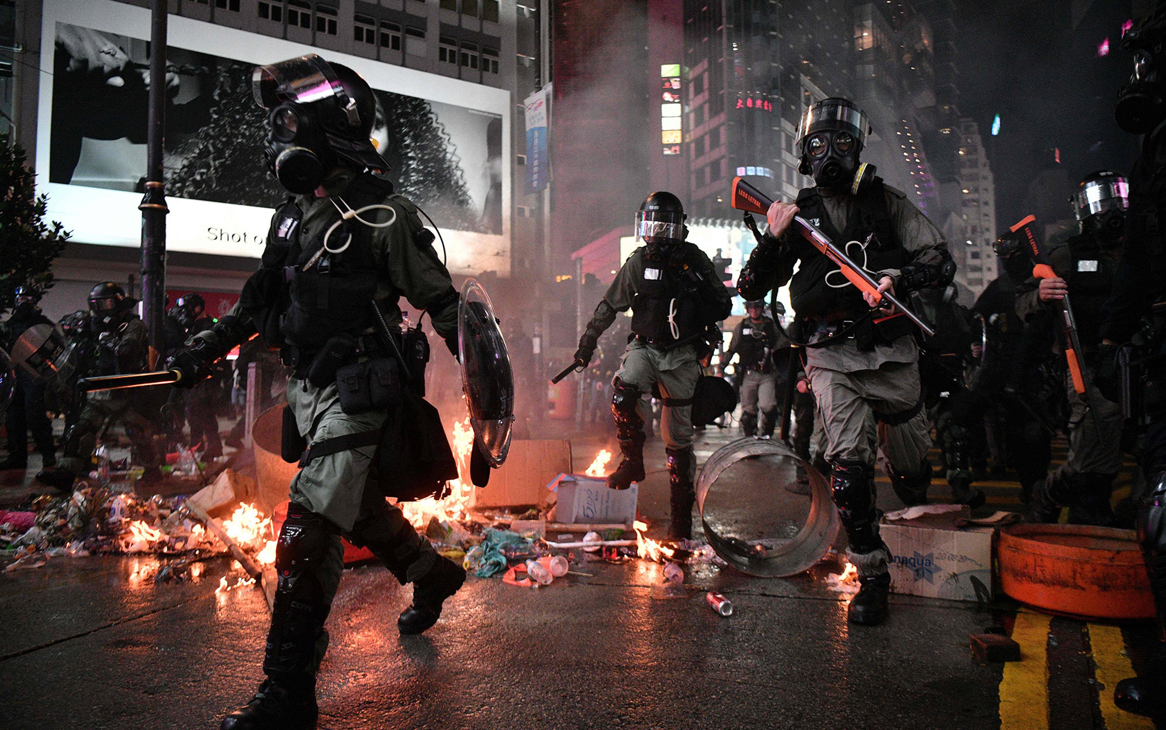 Riot police in gas masks carrying batons and shields walking through a street with flames and debris at night.