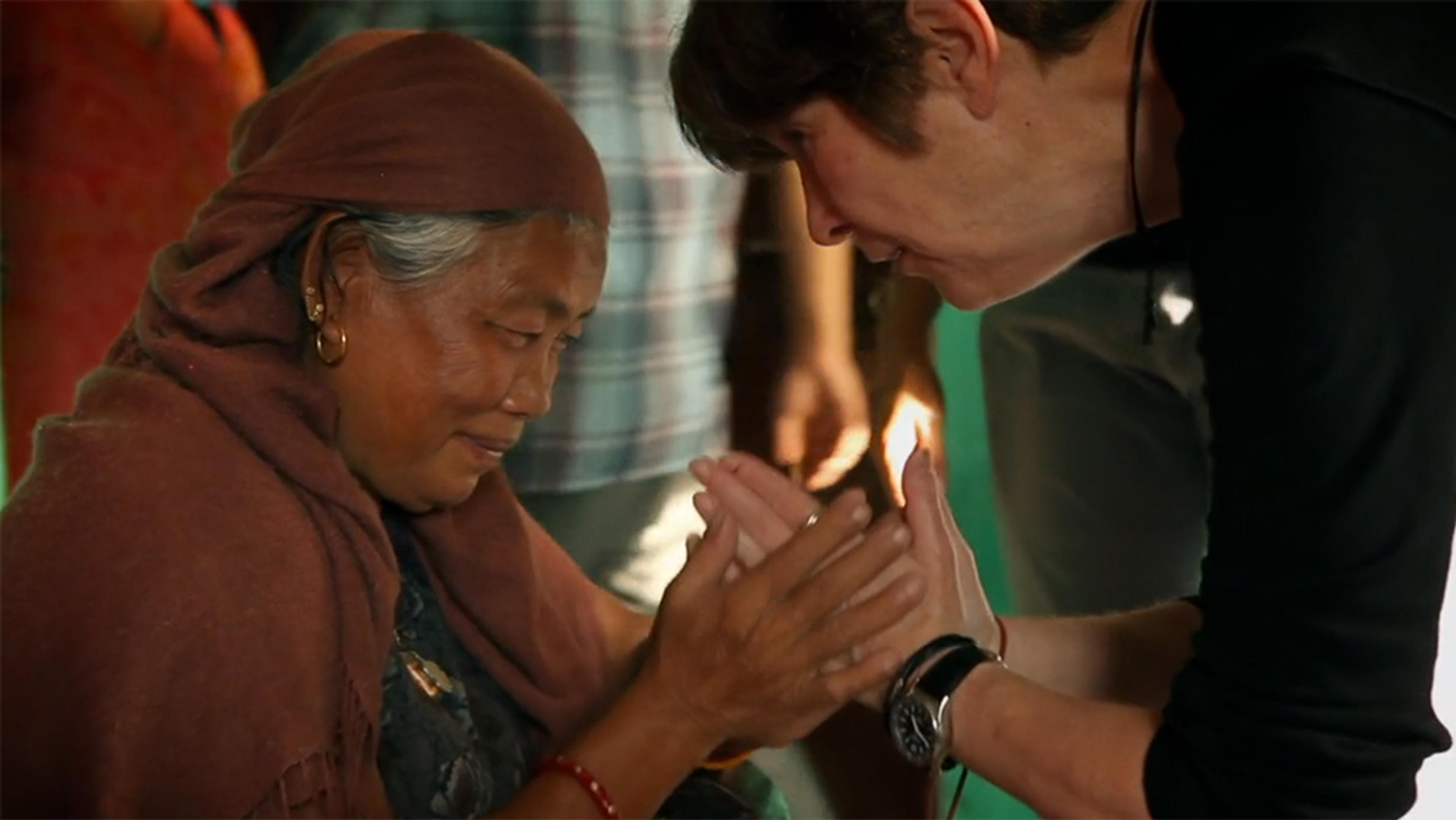An elderly Asian woman in a headscarf touches hands with a white woman, sharing an emotional moment indoors.