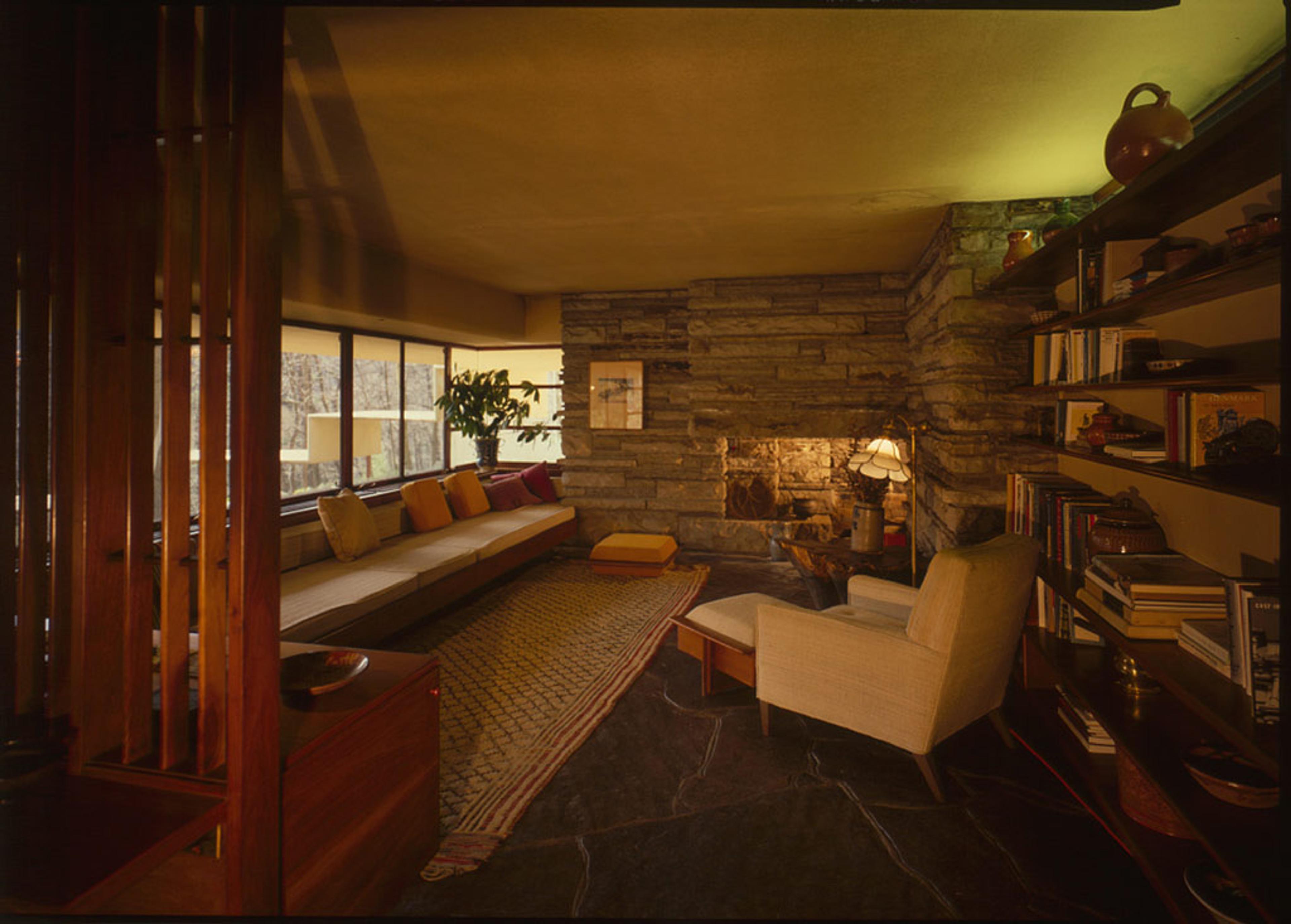 Photo of a cosy living room with a stone fireplace, built-in sofa with cushions, bookshelf and armchair. Warm lighting creates ambience.