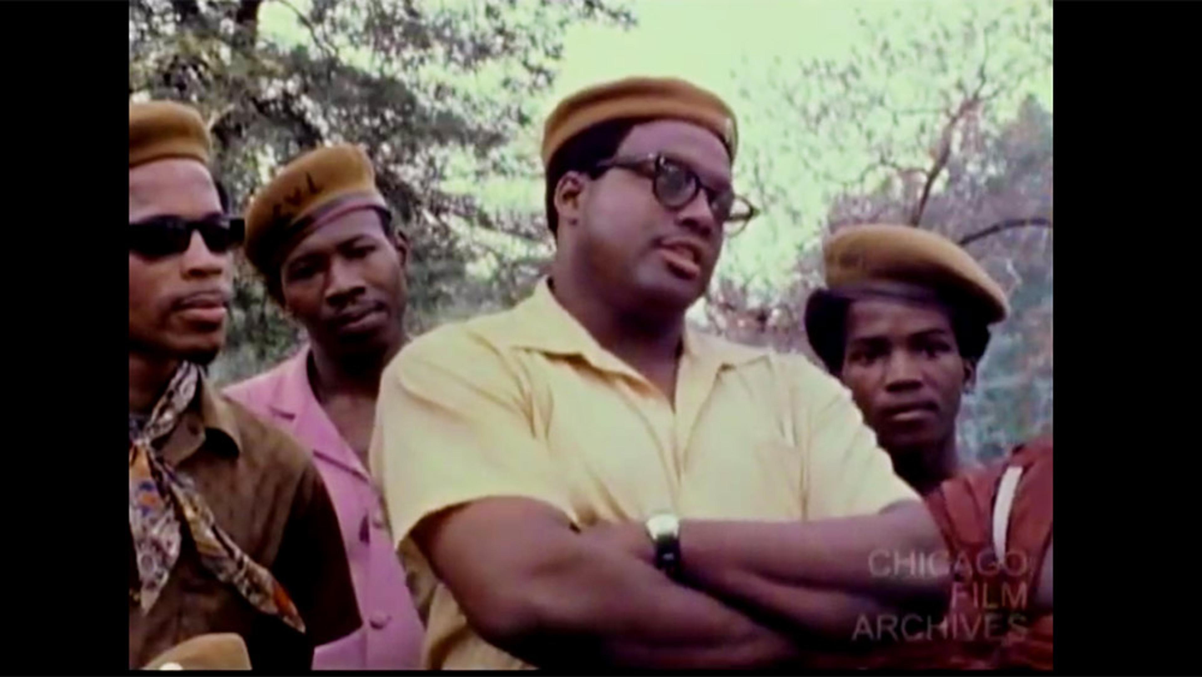 A vintage photo of a group of Black men wearing similar outfits, including yellow berets, standing outdoors with trees in the background.