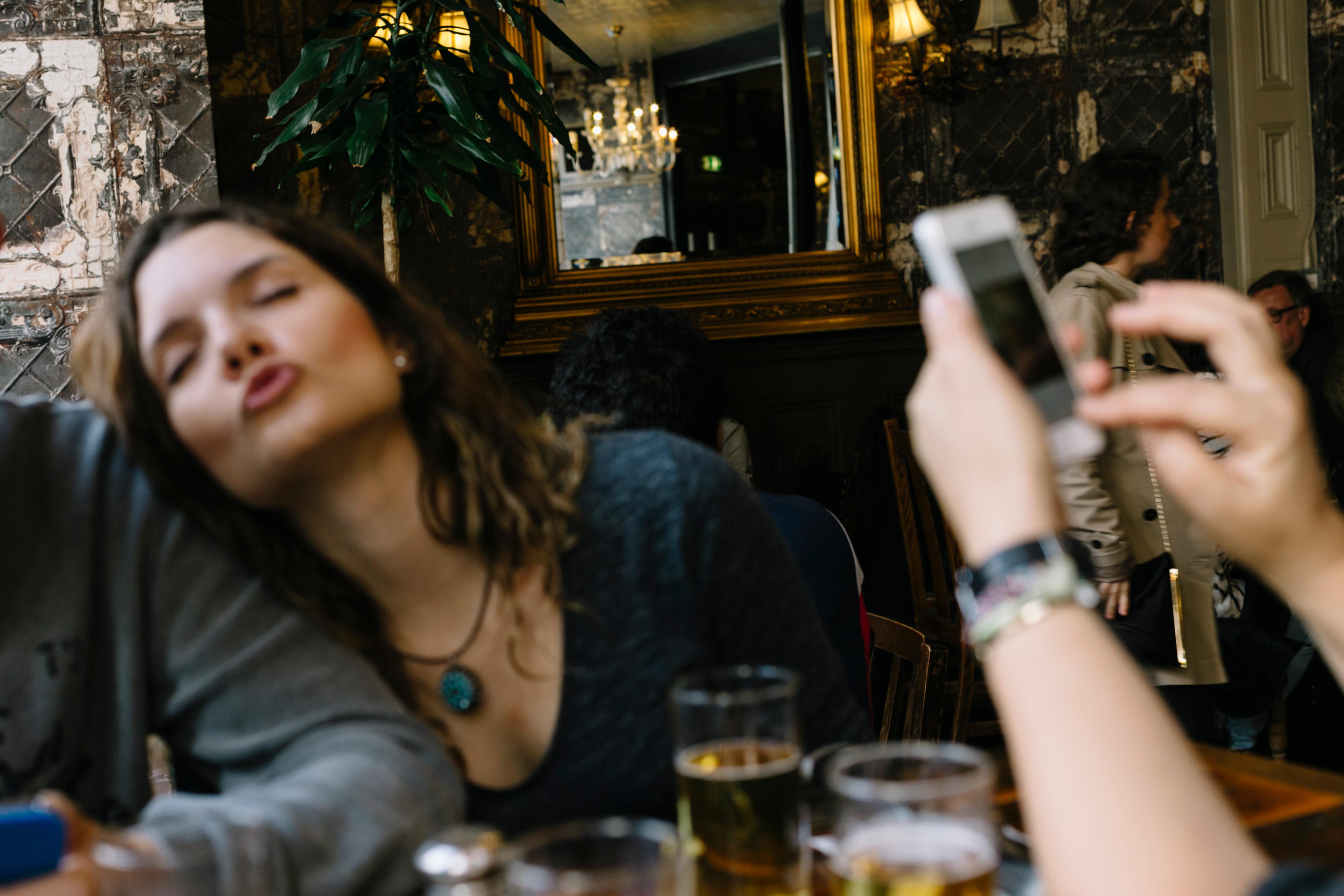A photo of a woman puckering her lips in a pub setting with drinks on the table and a hand holding a smartphone.