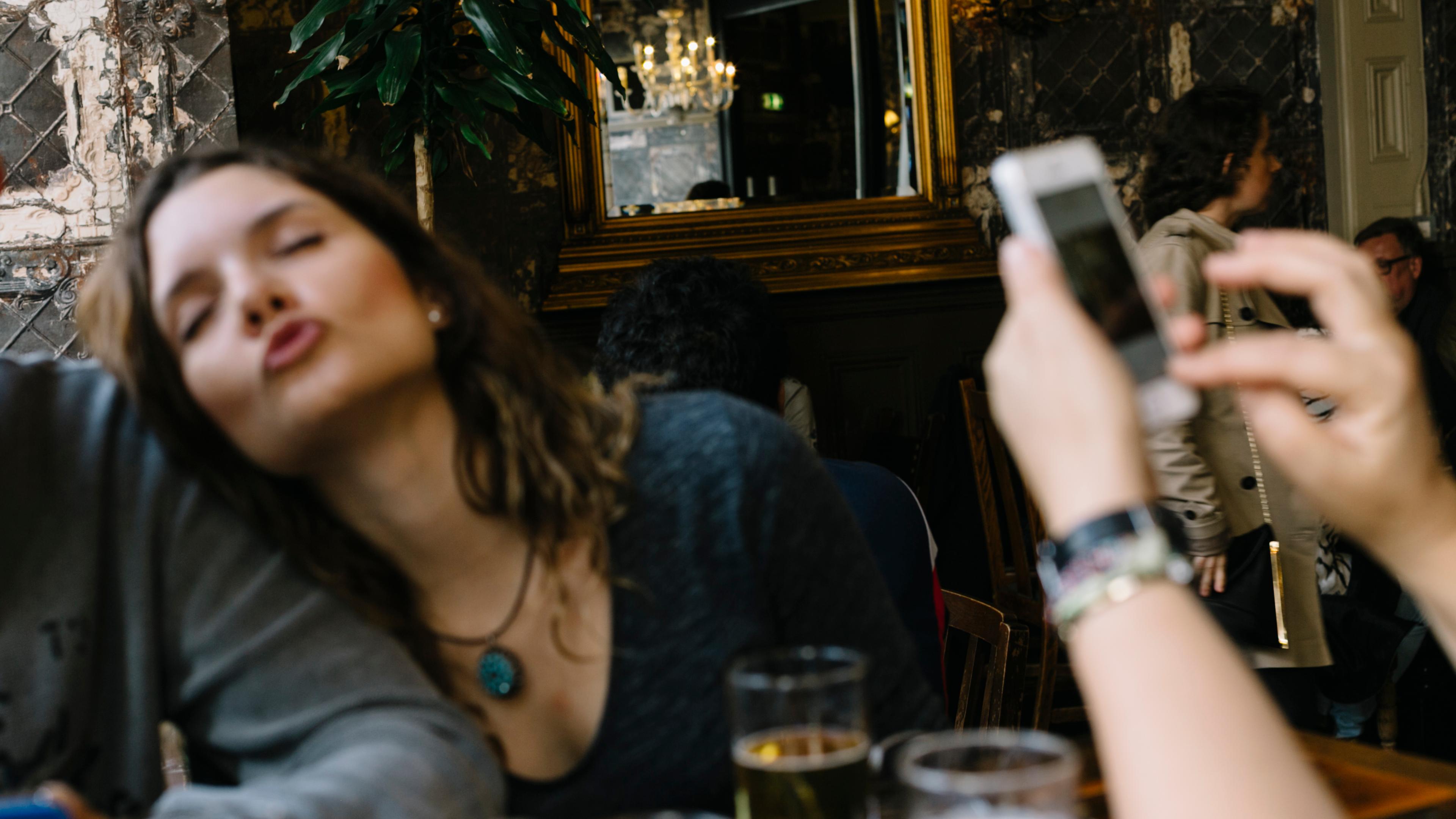 A photo of a woman puckering her lips in a pub setting with drinks on the table and a hand holding a smartphone.