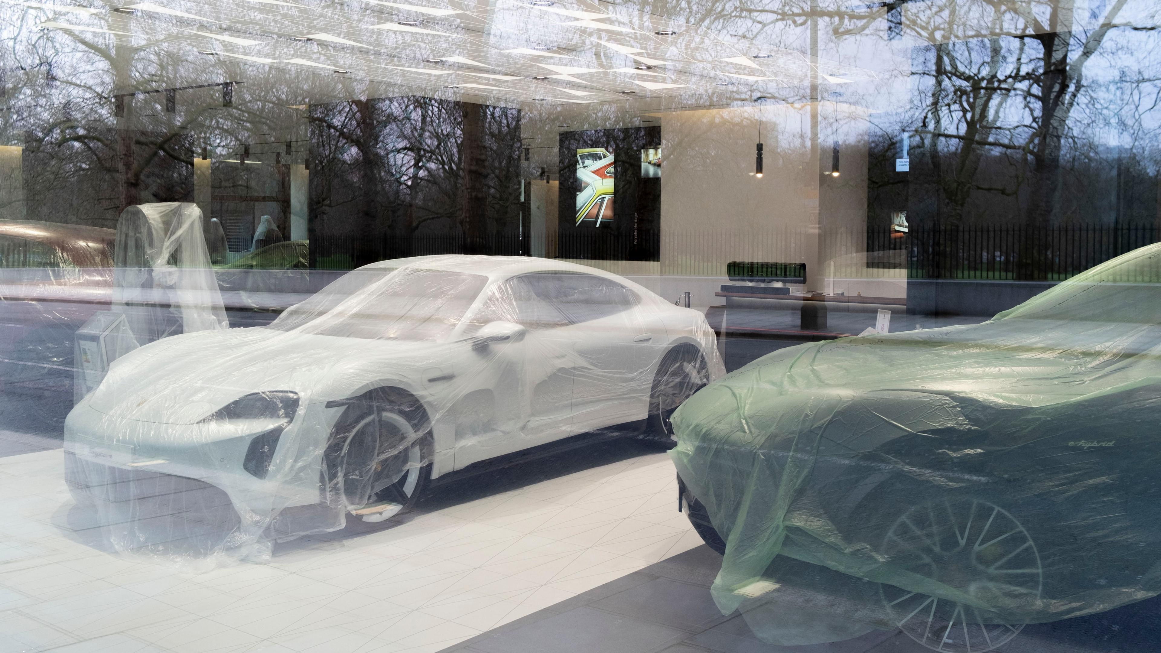 Photo of cars covered in protective plastic inside a showroom with reflections of bare trees in the glass facade.