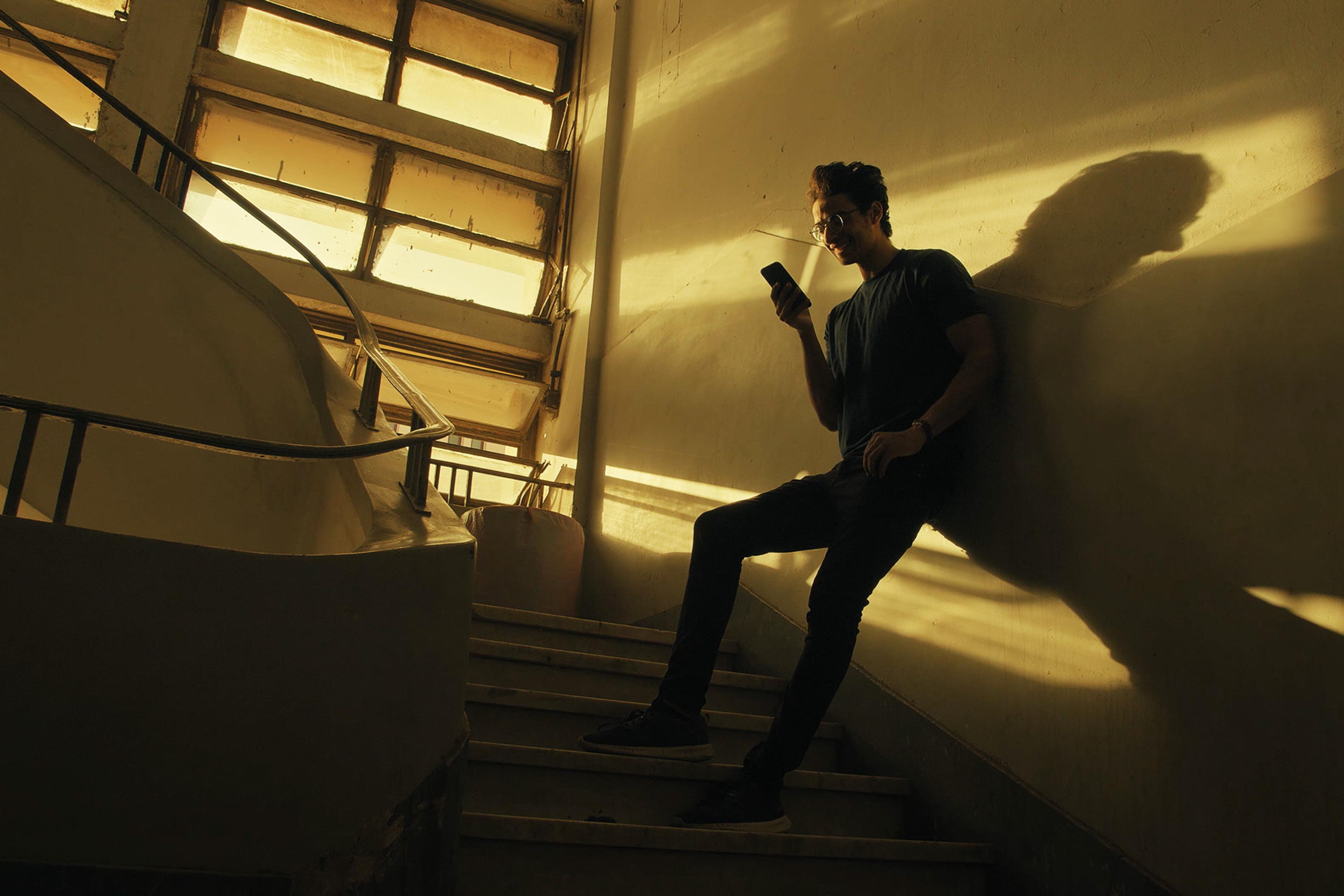 A person stands on a staircase in dim light, looking at a phone. Sunlight streams through large windows, casting shadows on the walls and stairs.