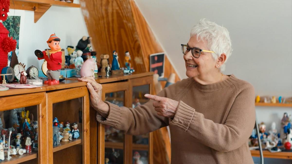 Photo of a smiling woman with short white hair pointing at a doll on a wooden cabinet filled with various figurines.