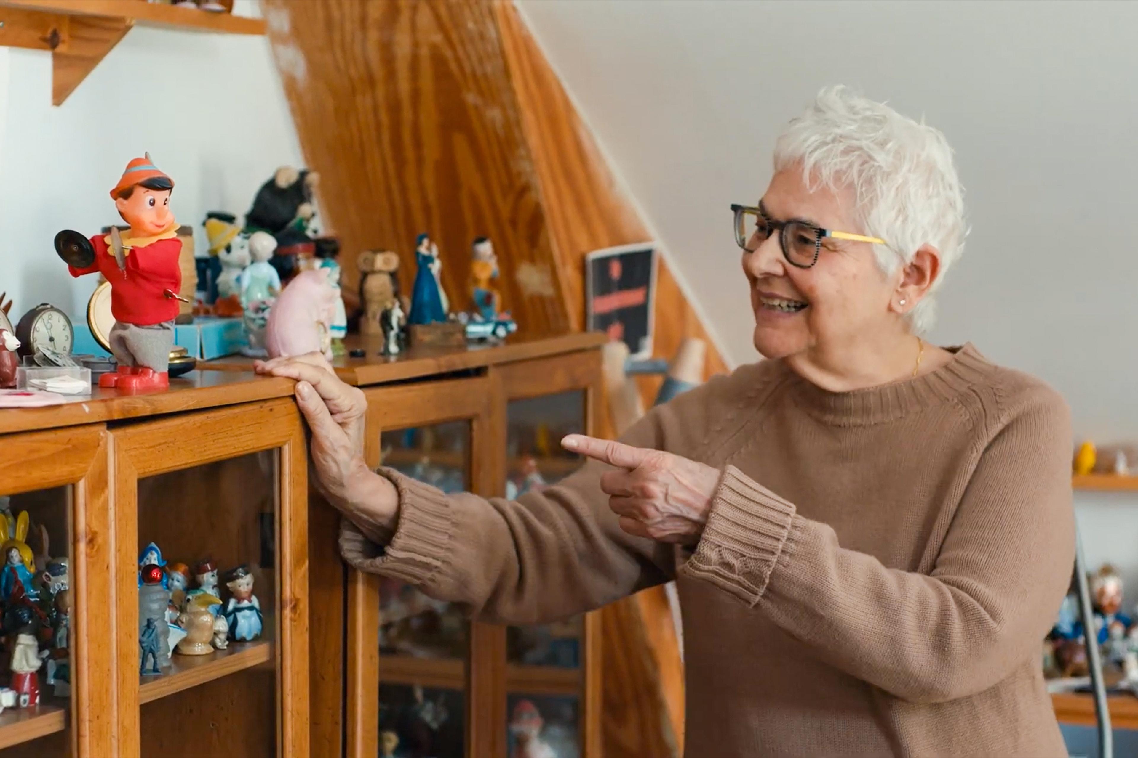 Photo of a smiling woman with short white hair pointing at a doll on a wooden cabinet filled with various figurines.