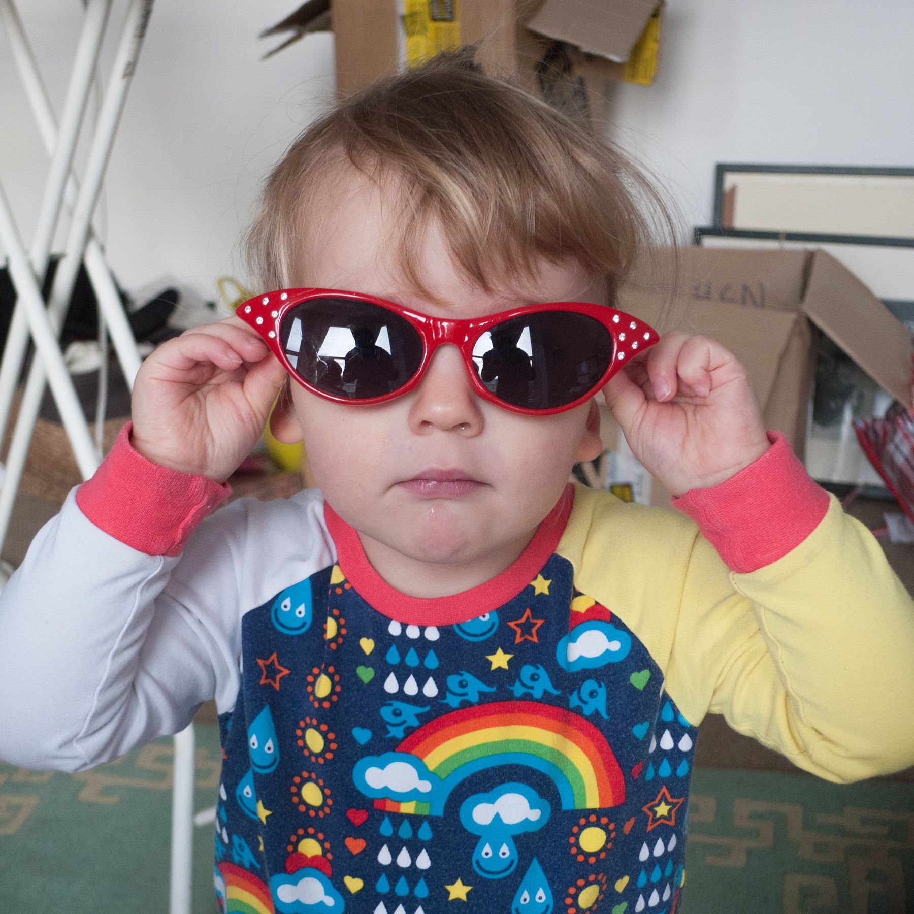 Photo of a child wearing red sunglasses and a colourful top with rainbows and clouds, standing indoors.
