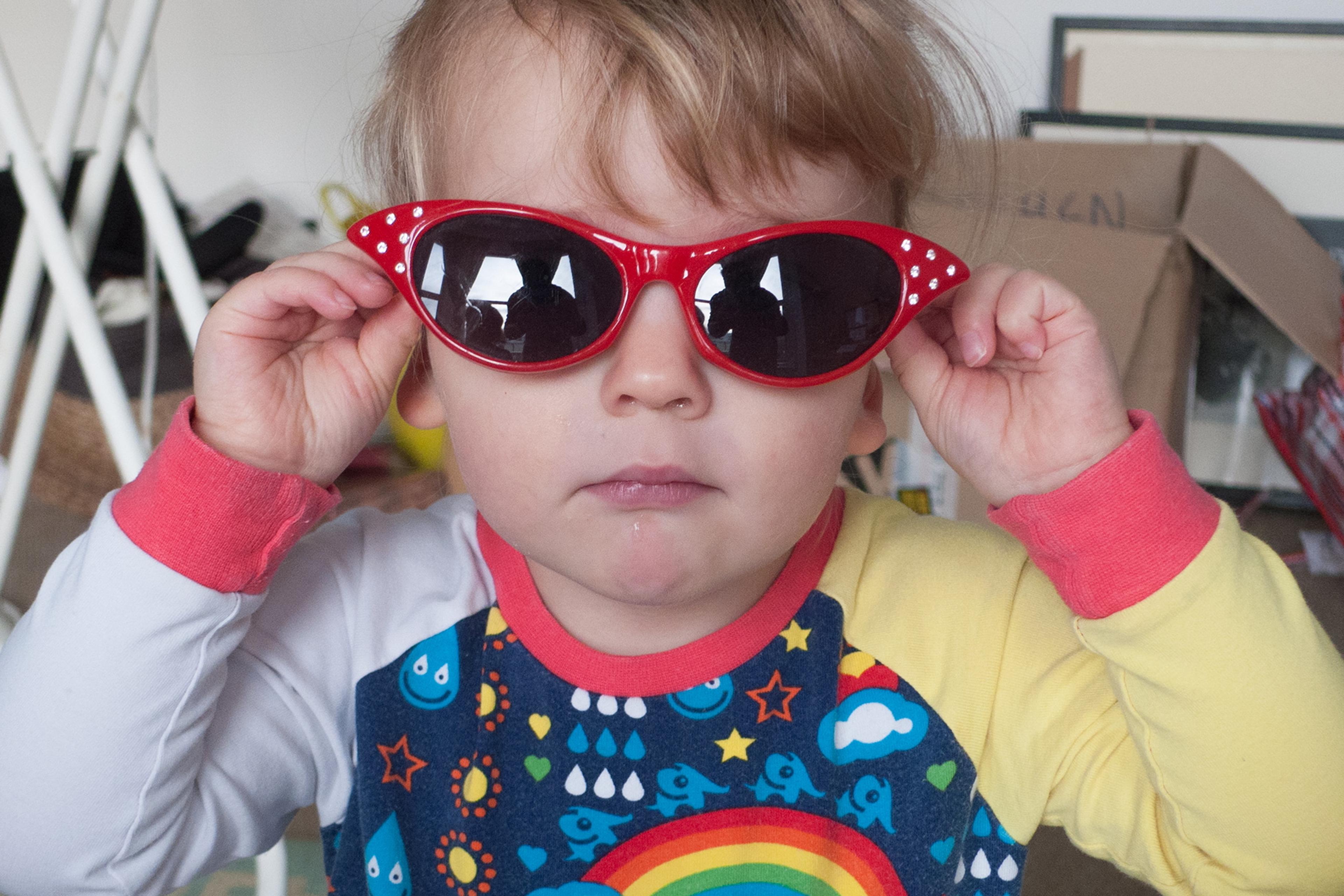 Photo of a child wearing red sunglasses and a colourful top with rainbows and clouds, standing indoors.