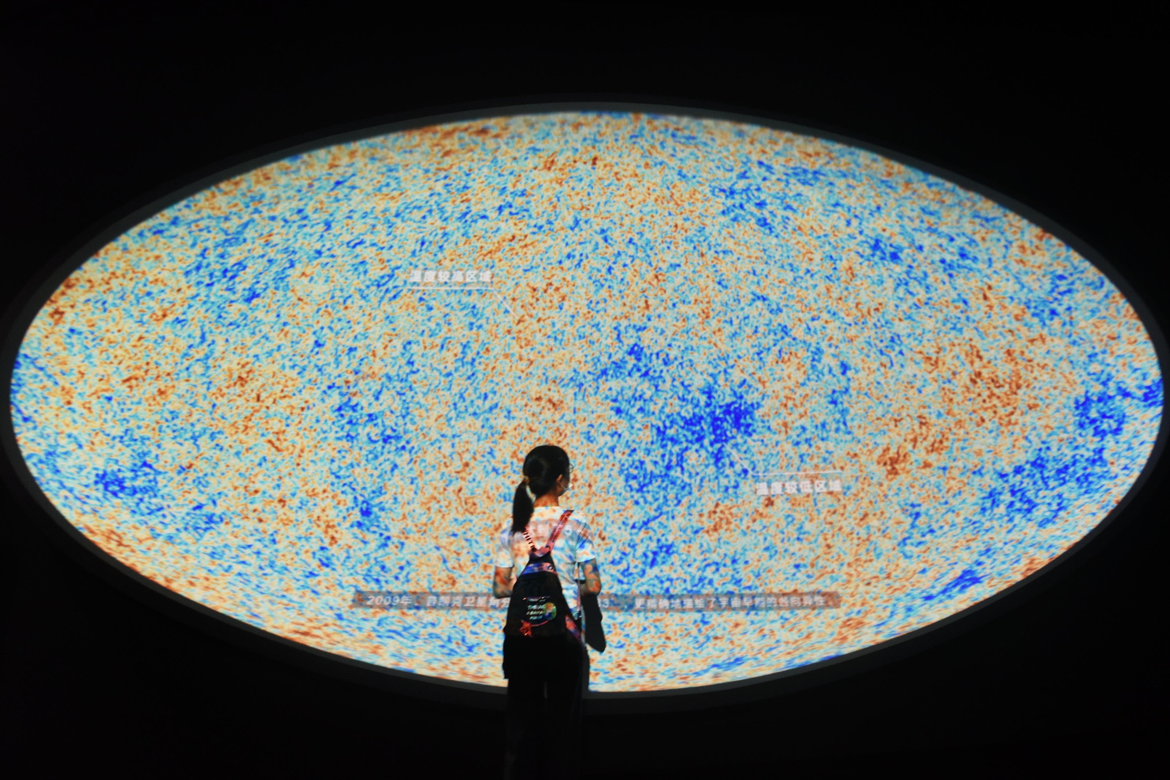 Photo of a person standing before a large, colourful cosmic map display with clumps of blue and orange speckles.