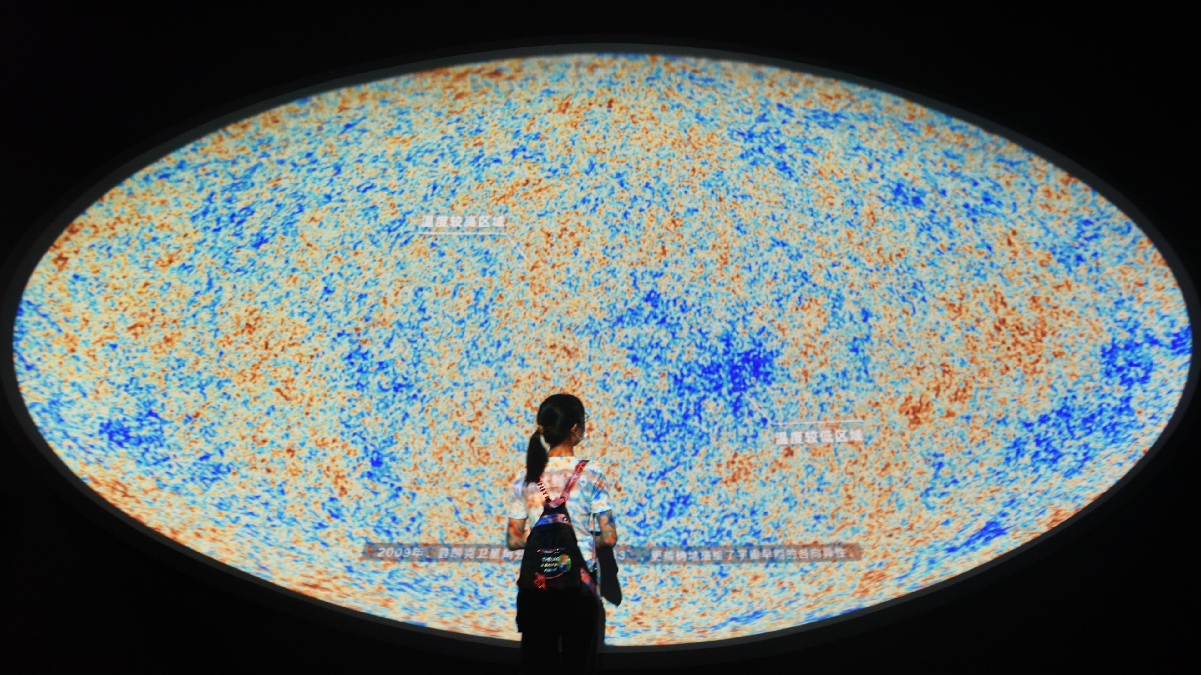 Photo of a person standing before a large, colourful cosmic map display with clumps of blue and orange speckles.