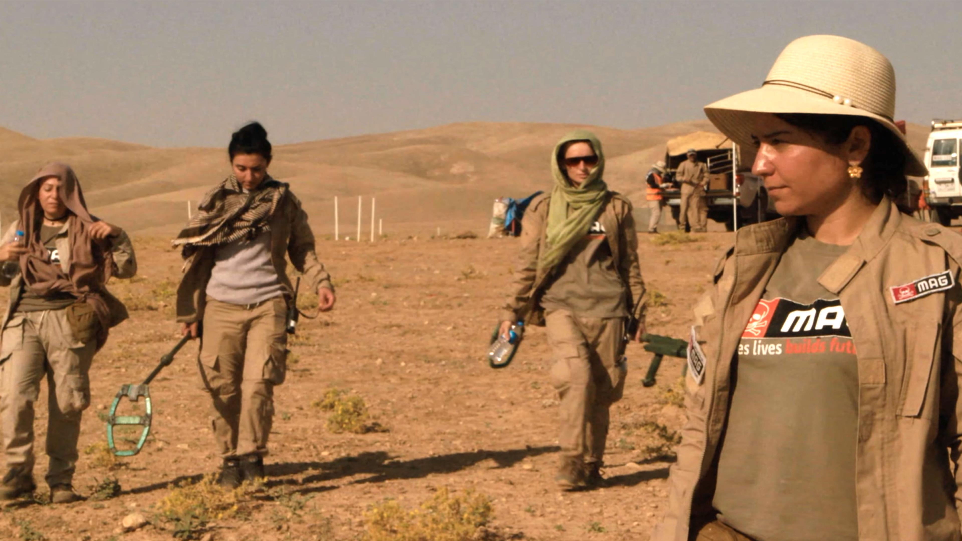 Photo of four women in desert gear walking across a dry landscape, carrying equipment with vehicles in the background.