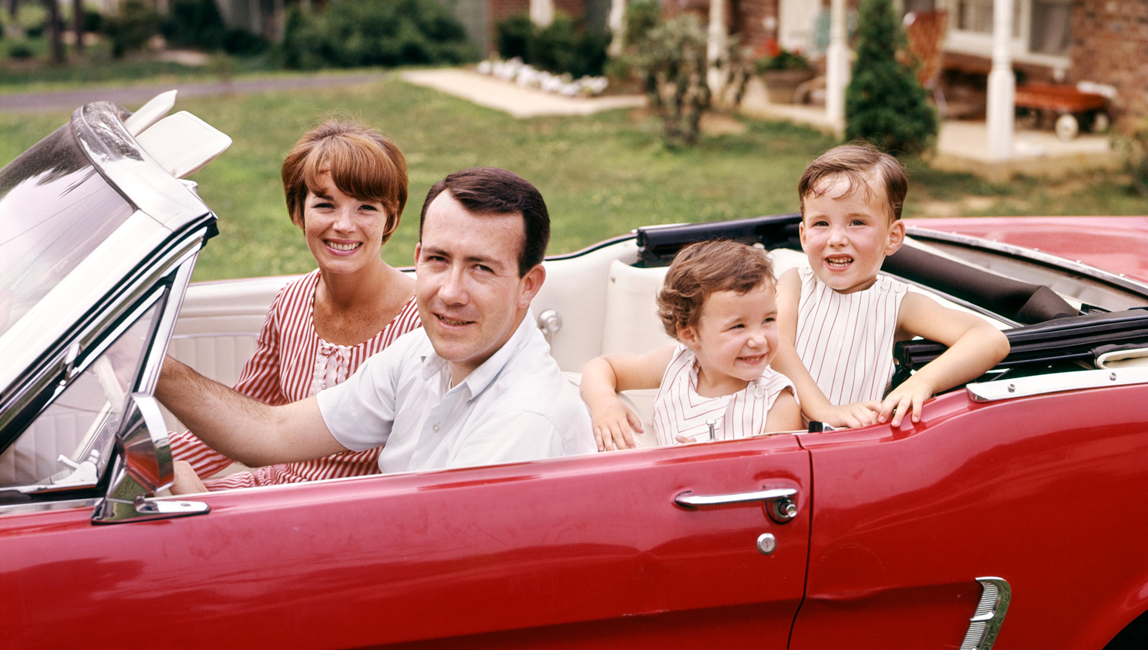 A family of four smiling and sitting in a red convertible car parked outside a house with green lawn.