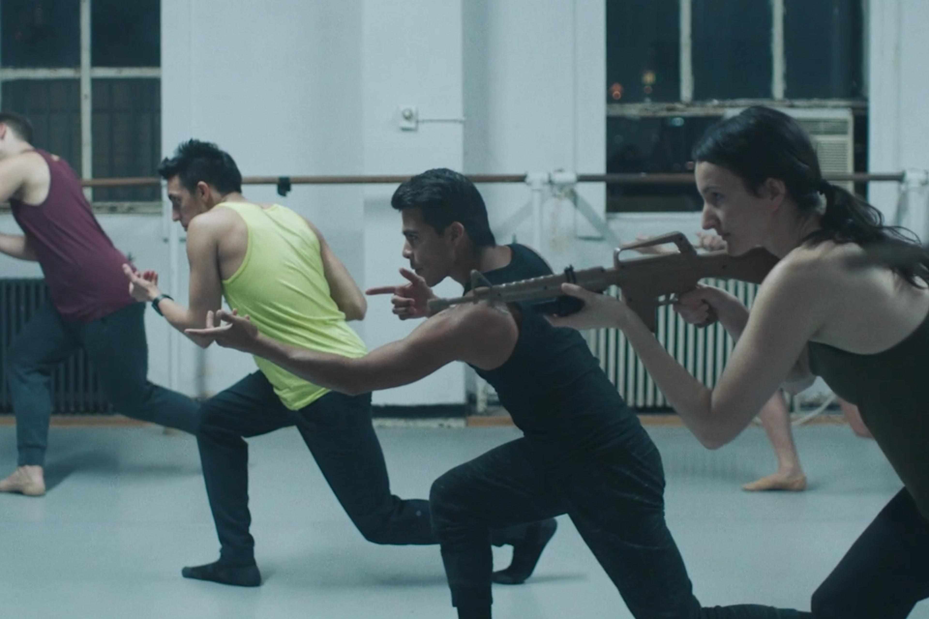 A group of dancers in a studio performing a dramatic routine, with one dancer holding a prop rifle aiming forward.