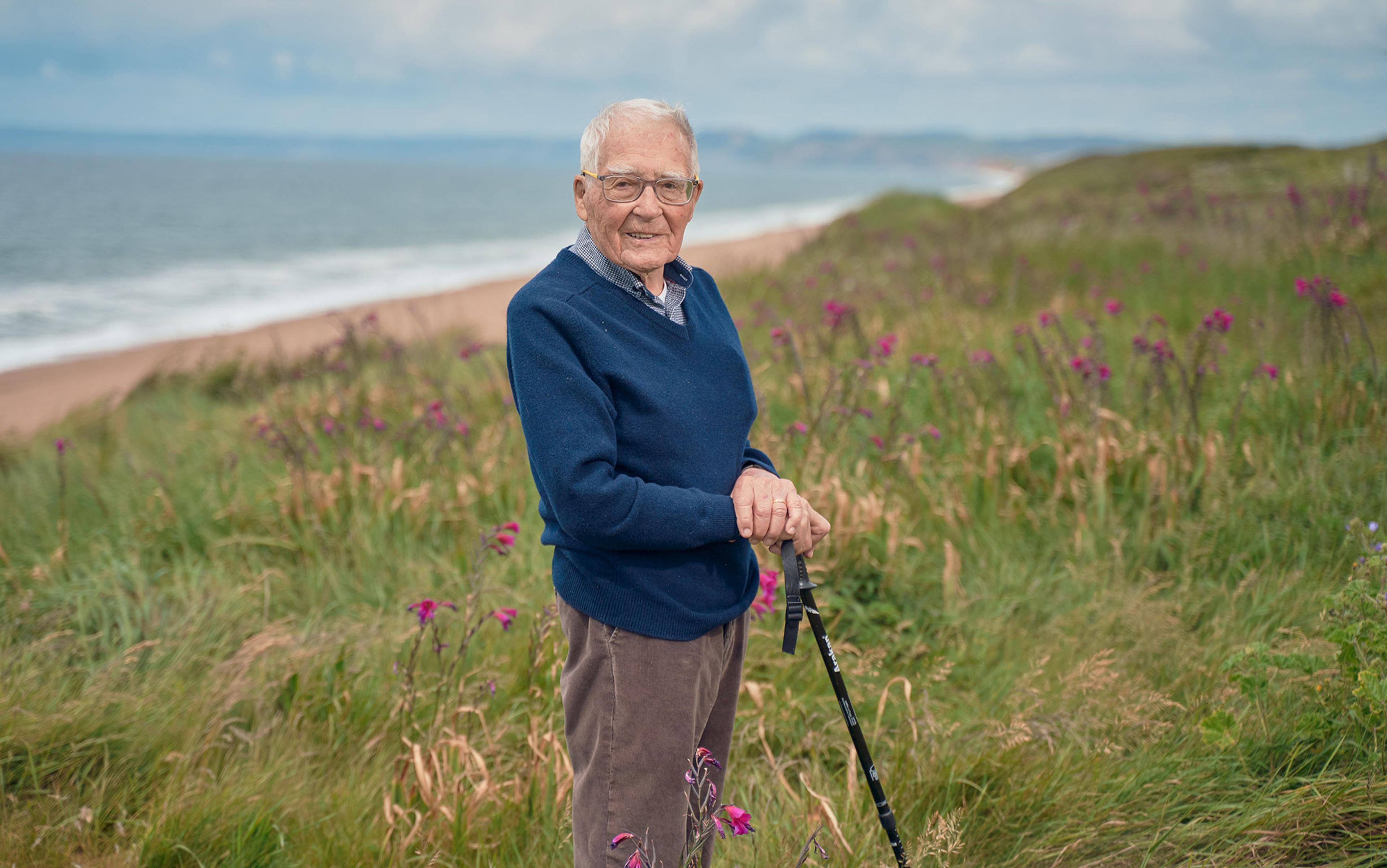 Elderly man with a walking stick stands smiling in a grassy field with wildflowers, near a beach and ocean under a cloudy sky.
