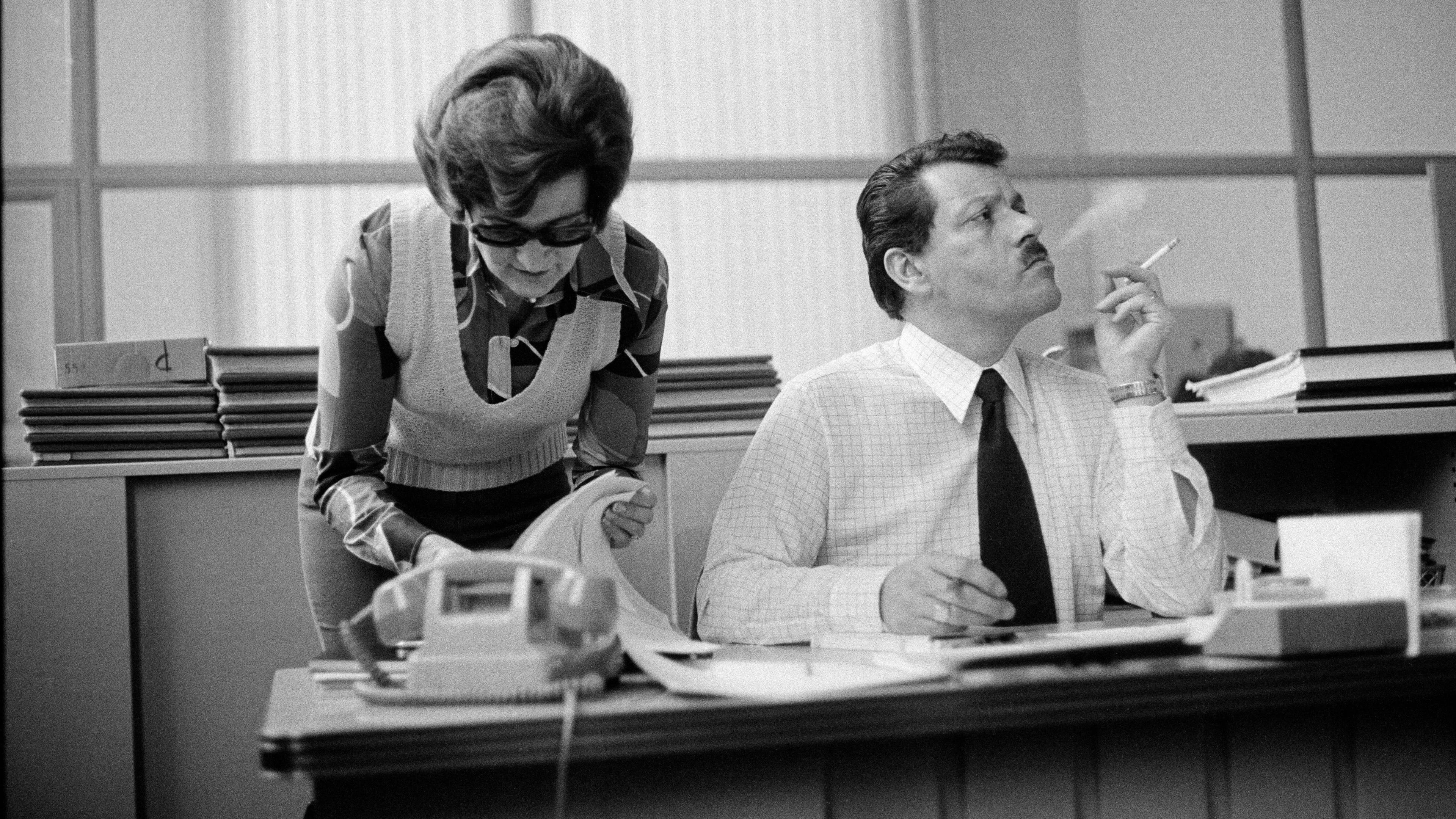 Black and white photo of a man smoking at a desk as a woman with glasses looks at papers beside him in an office setting.