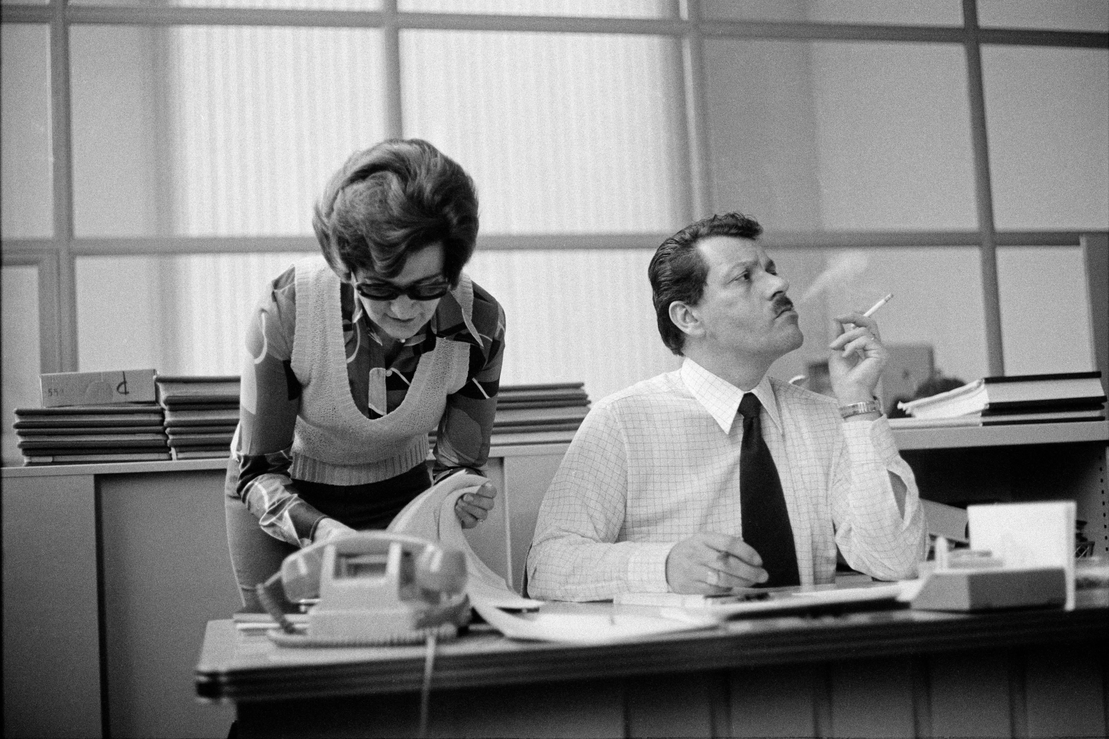 Black and white photo of a man smoking at a desk as a woman with glasses looks at papers beside him in an office setting.