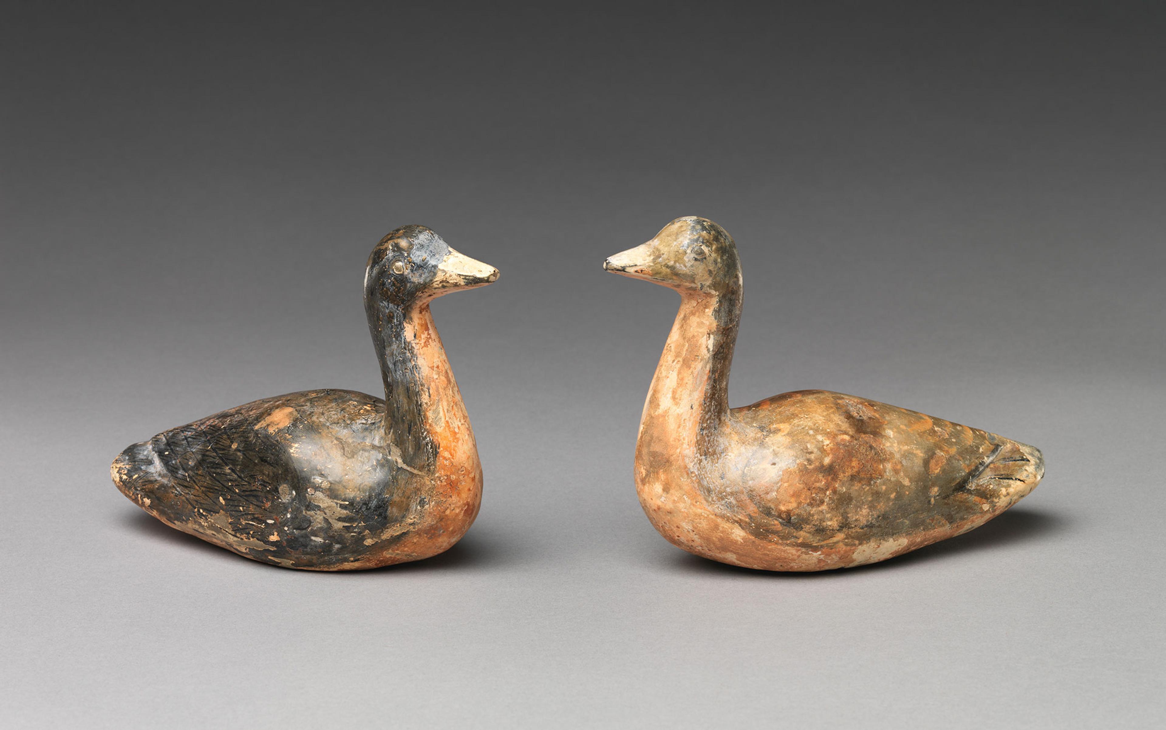 Two weathered, clay duck sculptures facing each other against a grey background.