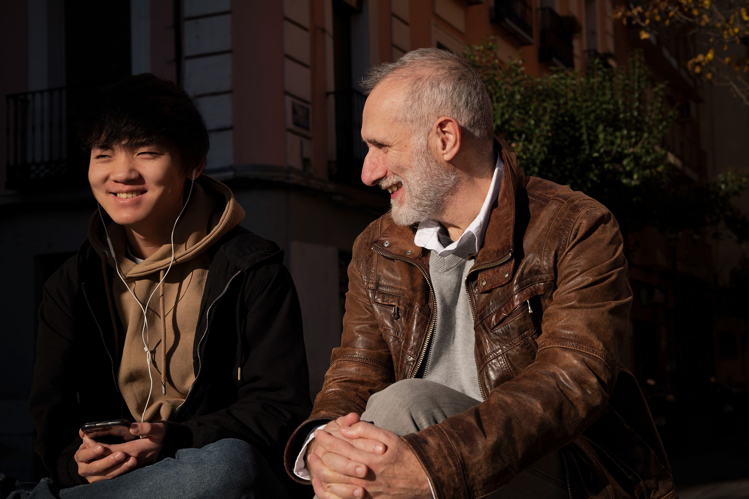 Two people sitting outdoors, one younger with headphones smiling, the other older looking at them while smiling, with trees in background.