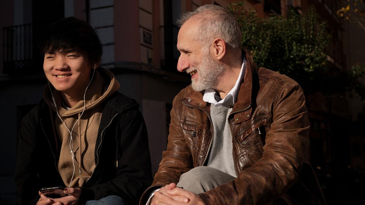 Two people sitting outdoors, one younger with headphones smiling, the other older looking at them while smiling, with trees in background.