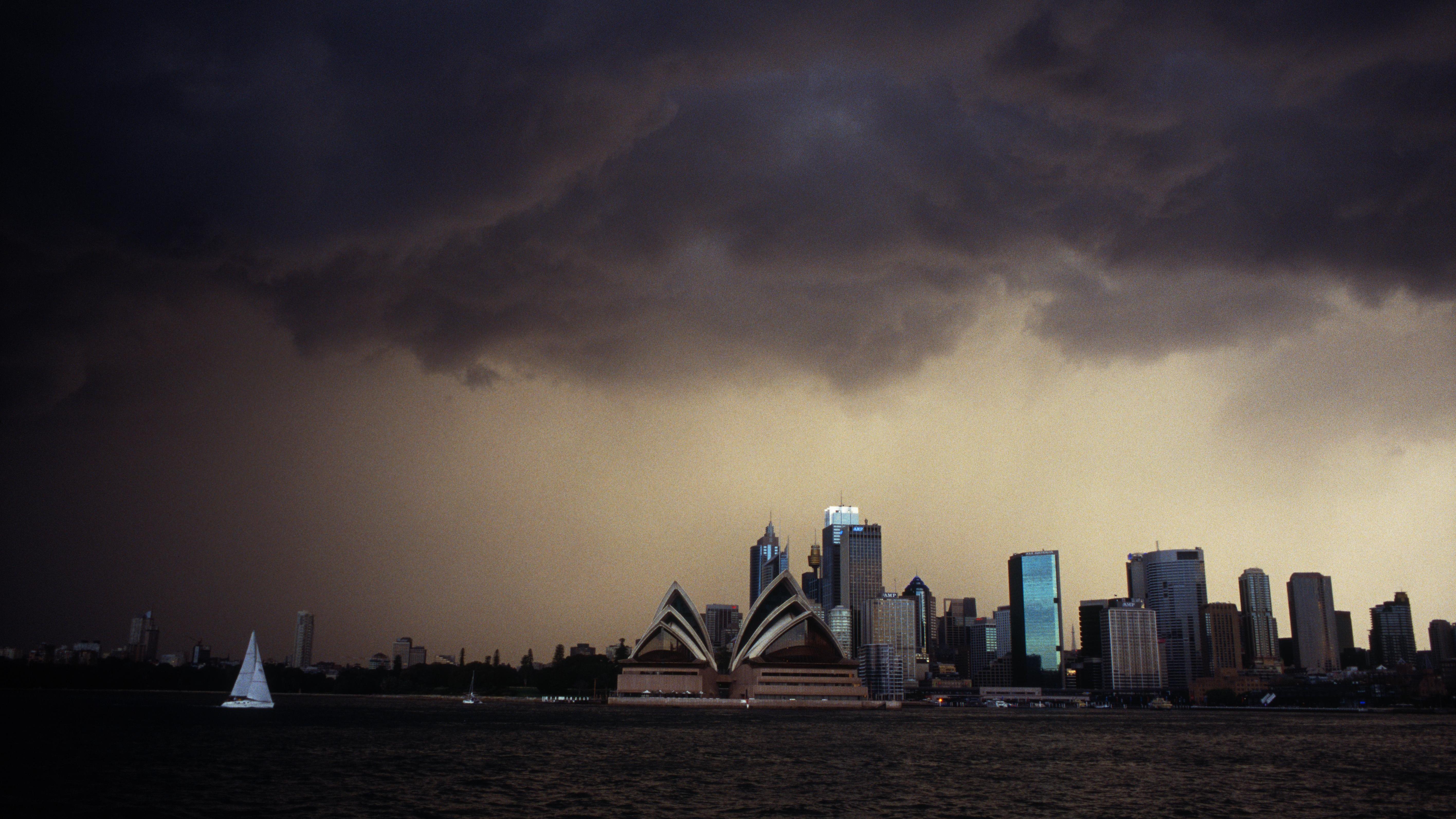 The Sydney city skyline featuring the Opera House under a dramatic dark stormy sky with a sailboat on the water in the foreground.