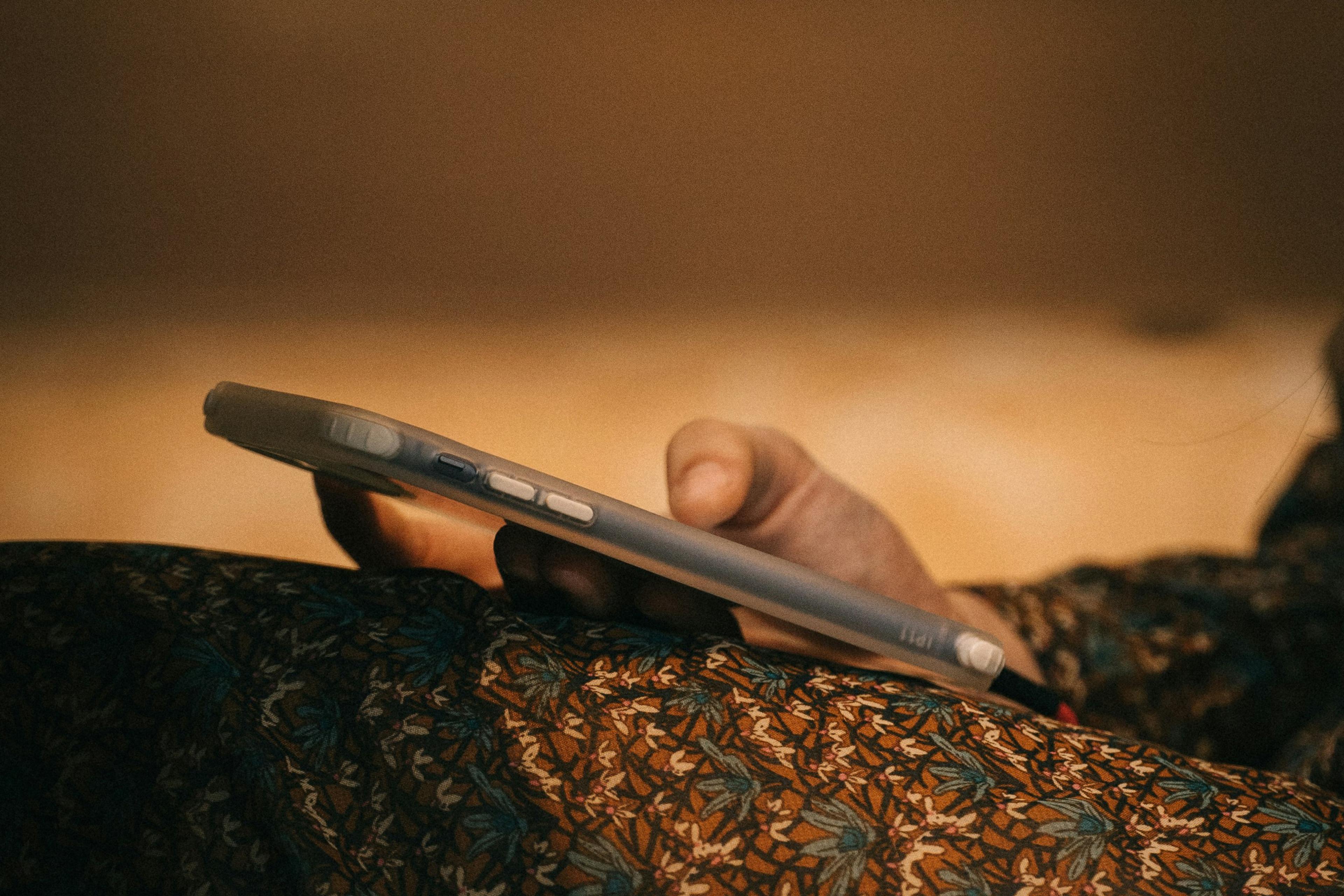 Close-up photo of a person’s hand holding a smartphone with a patterned fabric in the foreground, soft brown background.