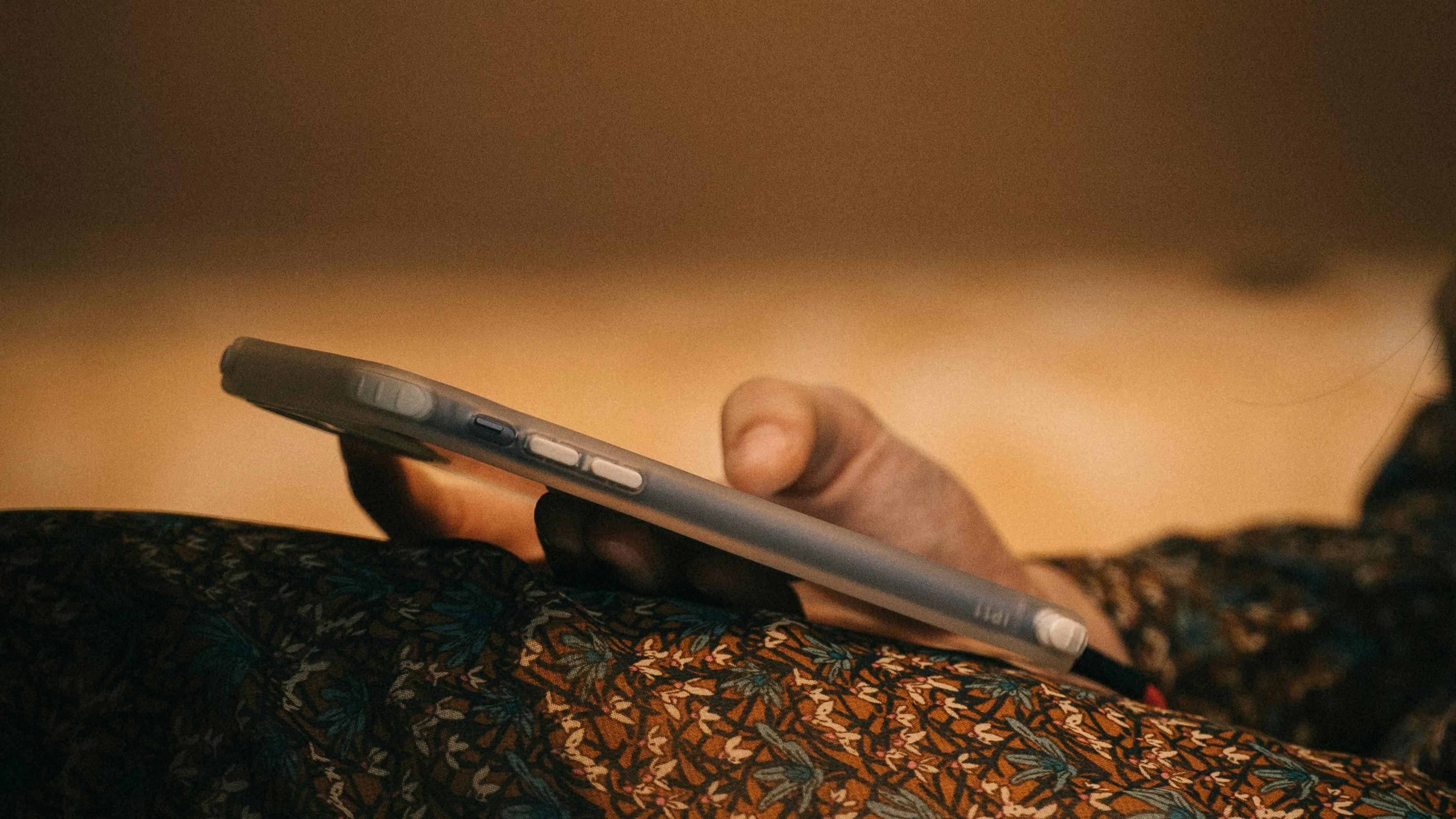 Close-up photo of a person’s hand holding a smartphone with a patterned fabric in the foreground, soft brown background.