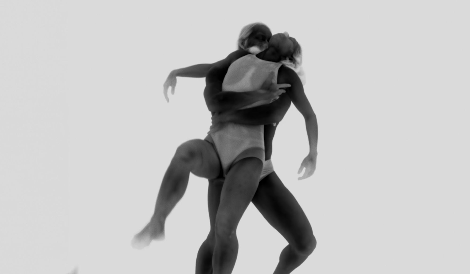 Black and white image of two dancers in swimsuits performing a dynamic lift, capturing the grace and movement against a plain background.