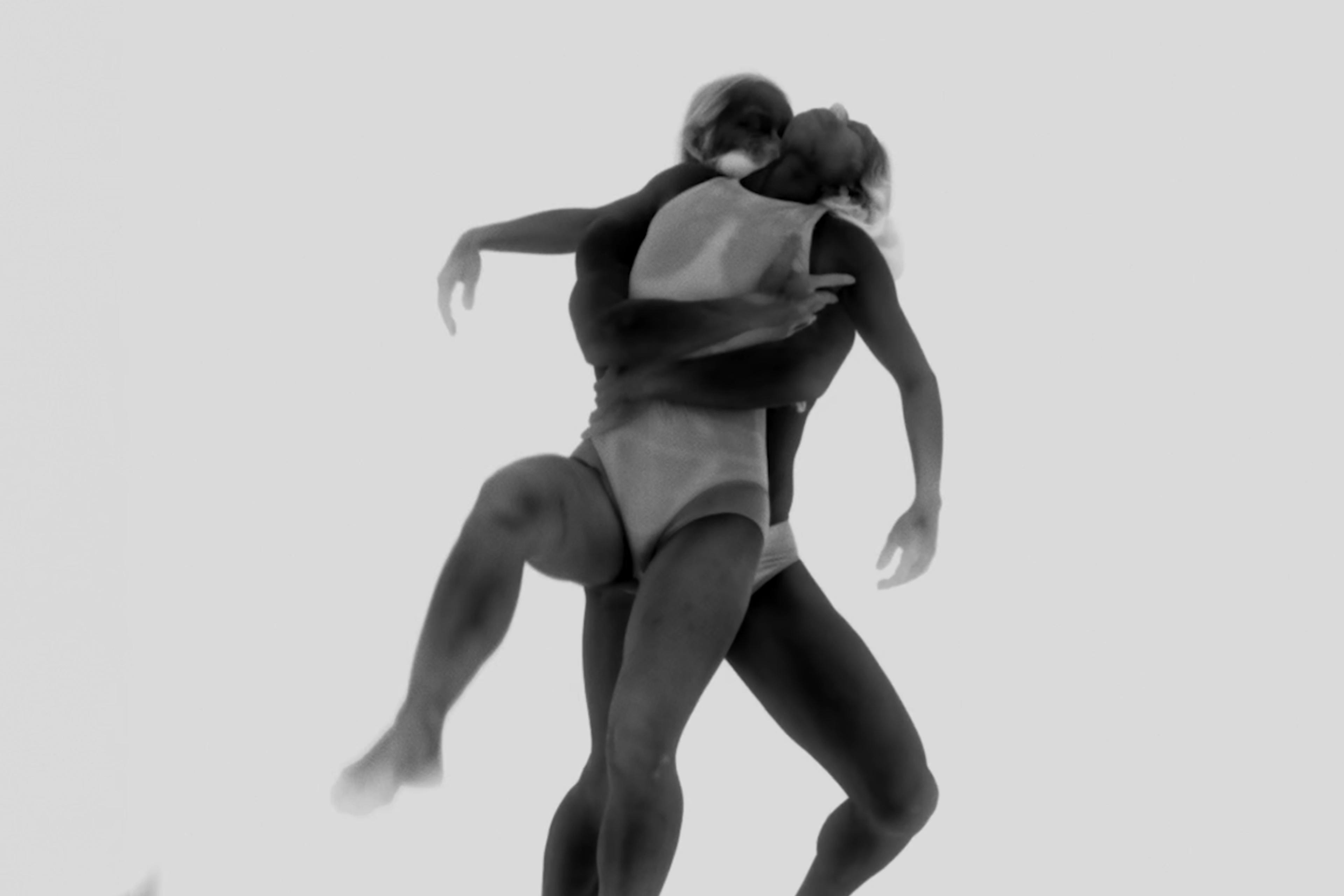 Black and white image of two dancers in swimsuits performing a dynamic lift, capturing the grace and movement against a plain background.