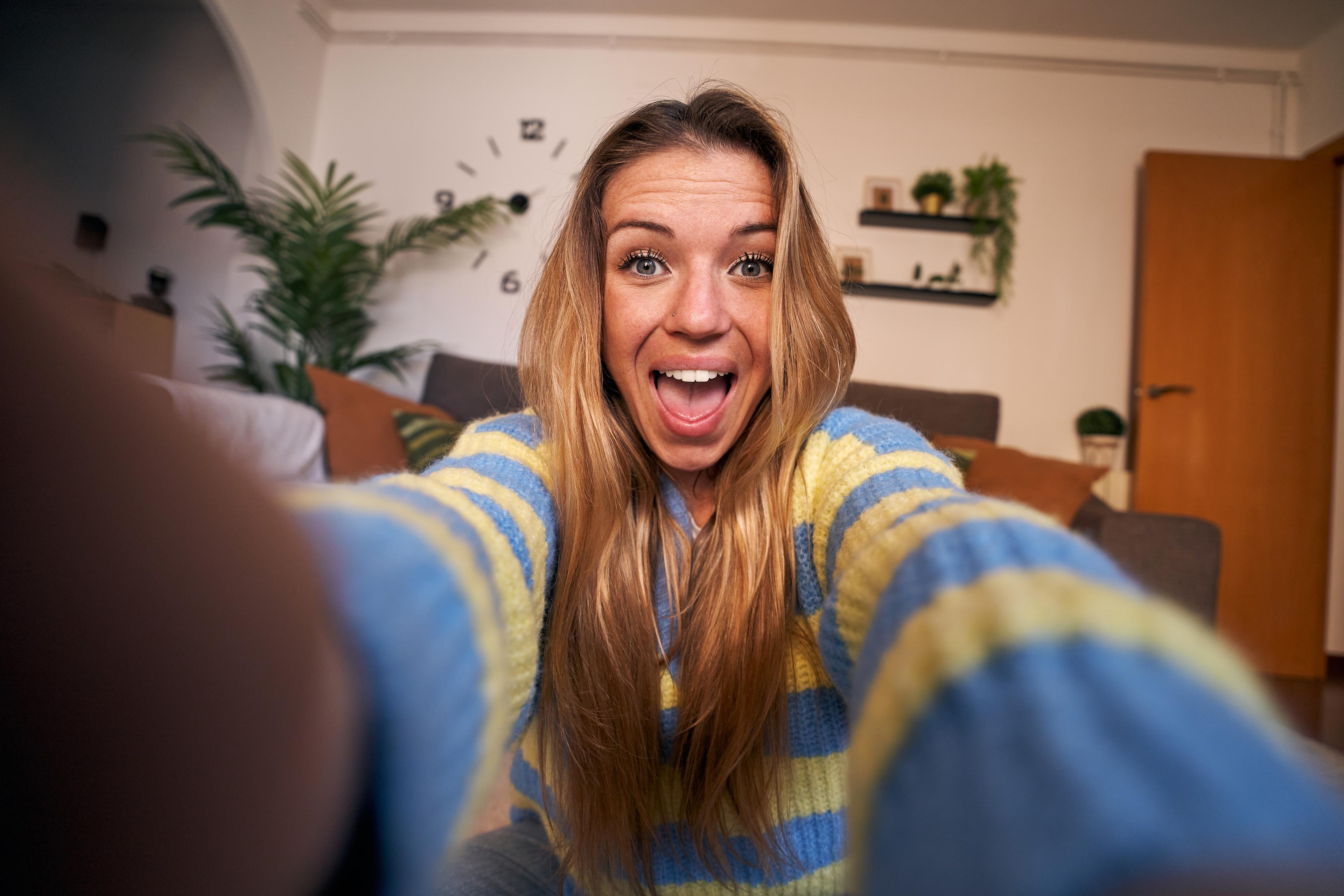Photo of a smiling person taking a selfie indoors, wearing a blue and yellow jumper, with plants and a clock in the background.