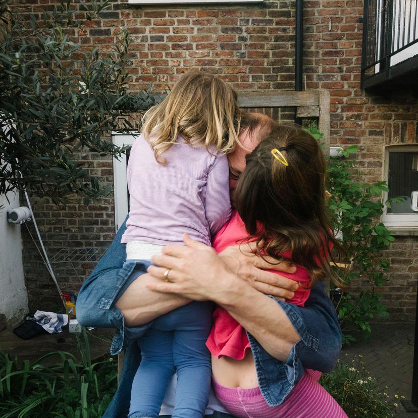 A person hugs two young children near a brick wall in a garden, with plants and greenery around them.
