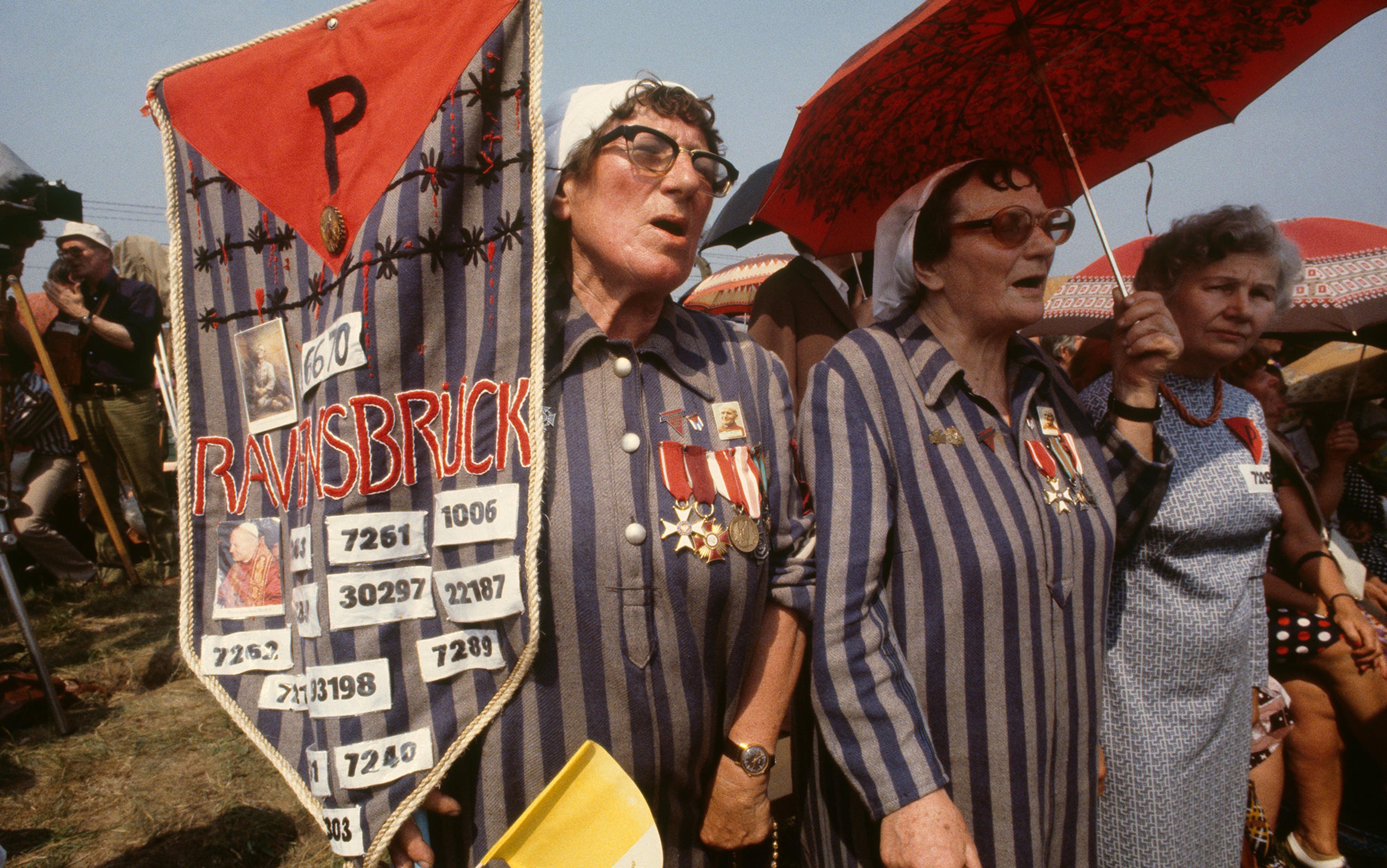 Women wearing striped uniforms and medals, holding a Ravensbrück banner and umbrellas, participating in a commemorative event outdoors.