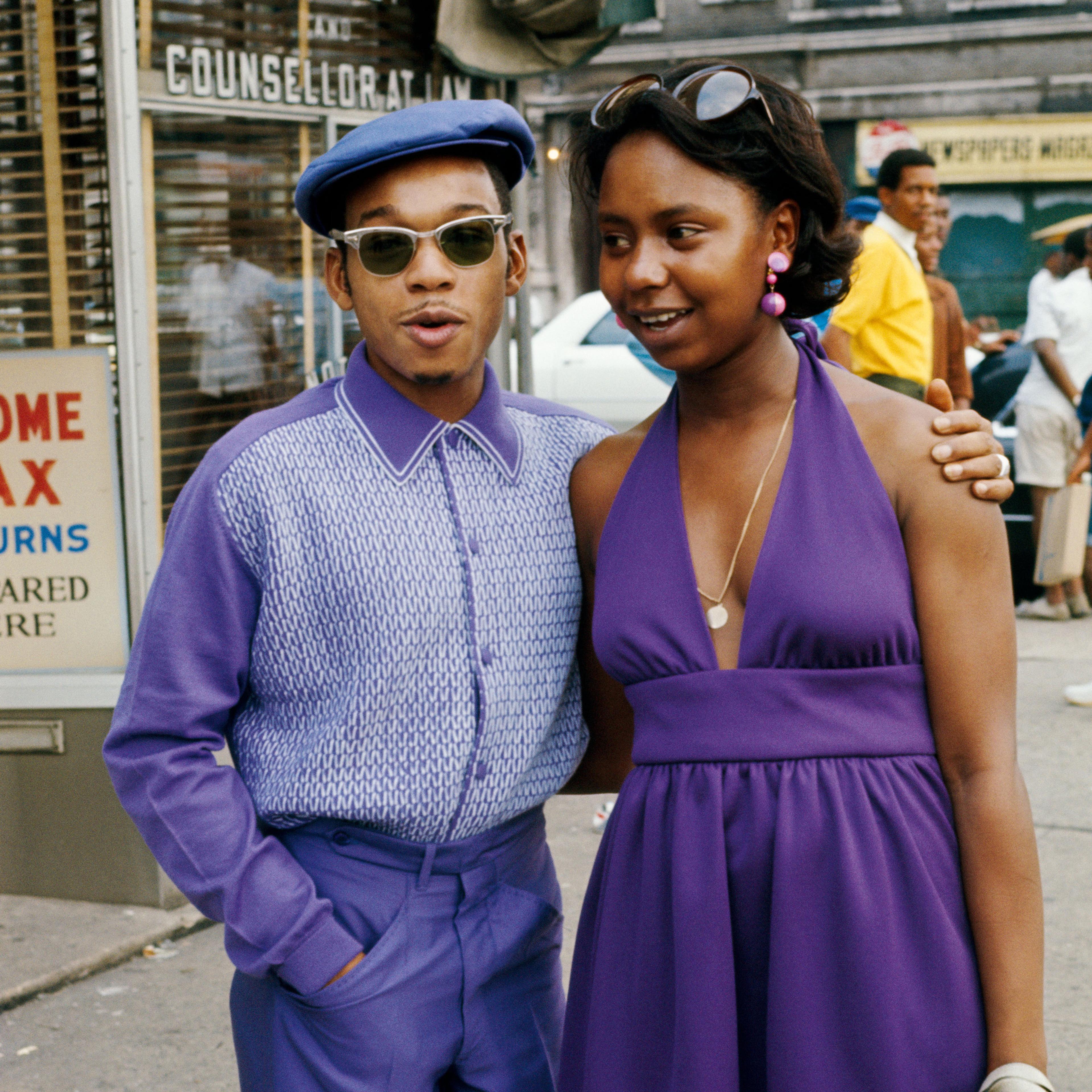 보라색 옷을 맞춰 입은 남녀가 도시 거리의 상점 쇼윈도 앞에서 미소를 지으며 포즈를 취하고 있다.<BR>