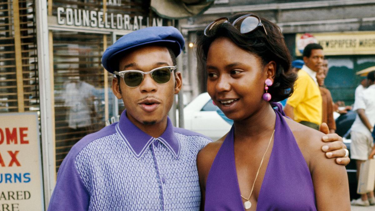 A man and woman in matching purple outfits on a city street, smiling and posing in front of a shop window.