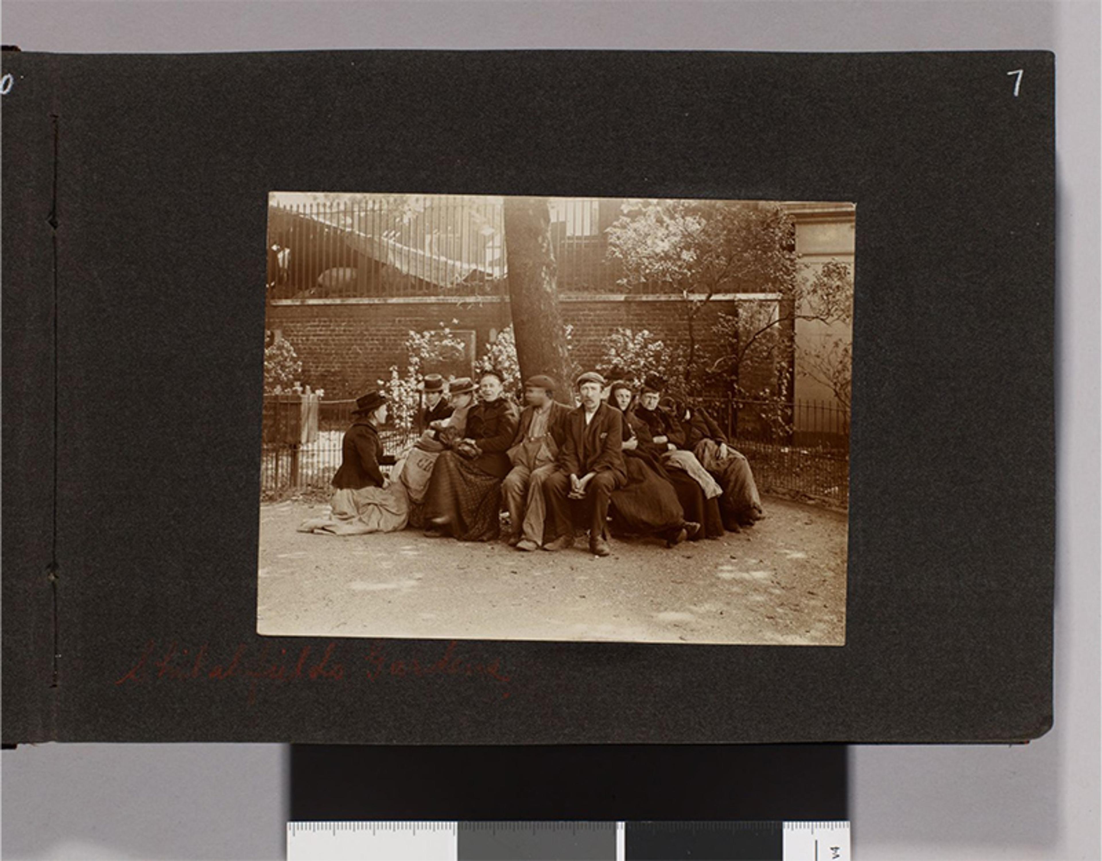 Black-and-white photo of people in Edwardian clothing sitting on a bench in a garden with a tree and brick building in the background.